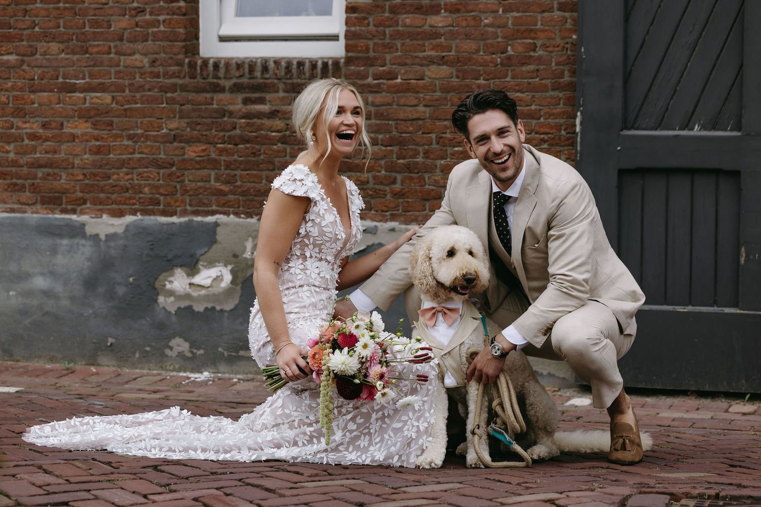 Couple poses with their white dog against a brick wall on their wedding day.