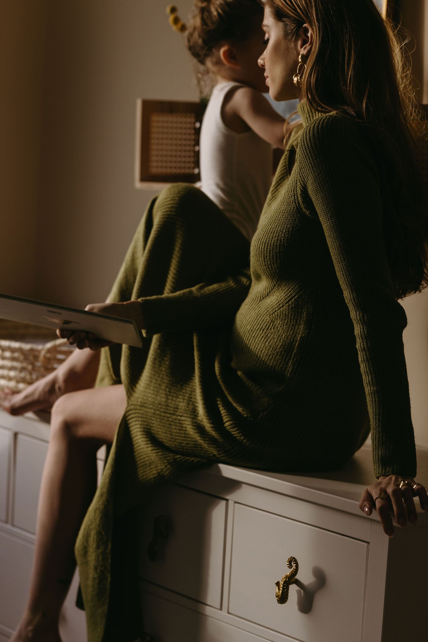 Woman in olive green sweater sitting on white furniture in moody lighting.