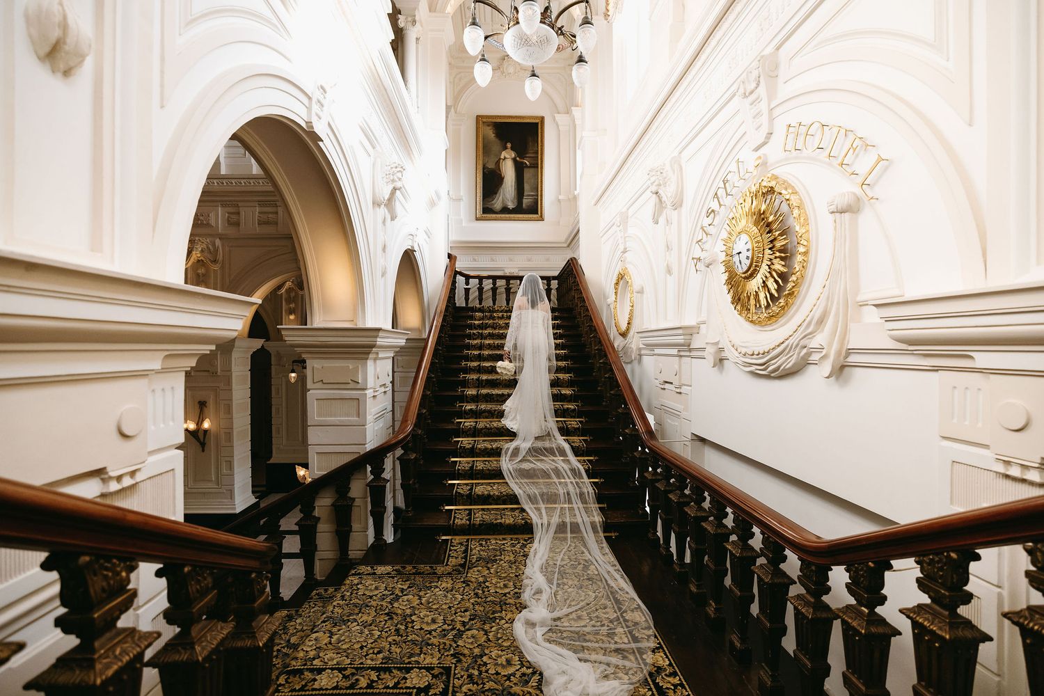 A grand white staircase with dark wood railings in an elegant baroque building featuring ornate architectural details and carpet.