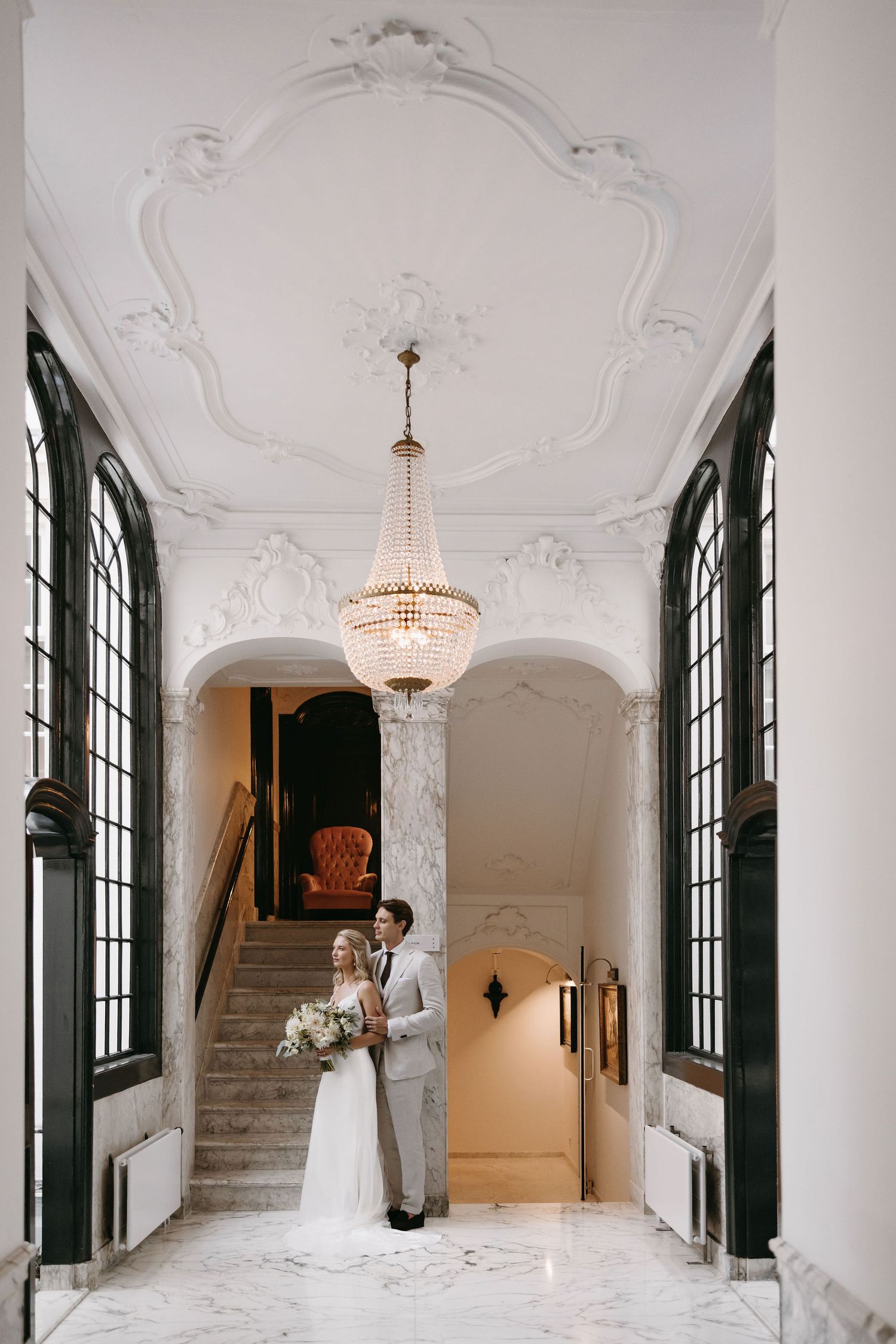 Two people stand beneath an ornate crystal chandelier in a grand white foyer with decorative moldings and tall windows.