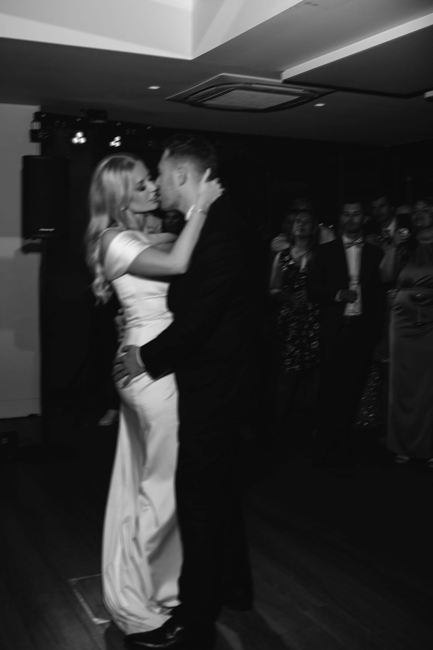 A couple shares a romantic dance at a wedding reception in black and white, with onlookers visible in the background.