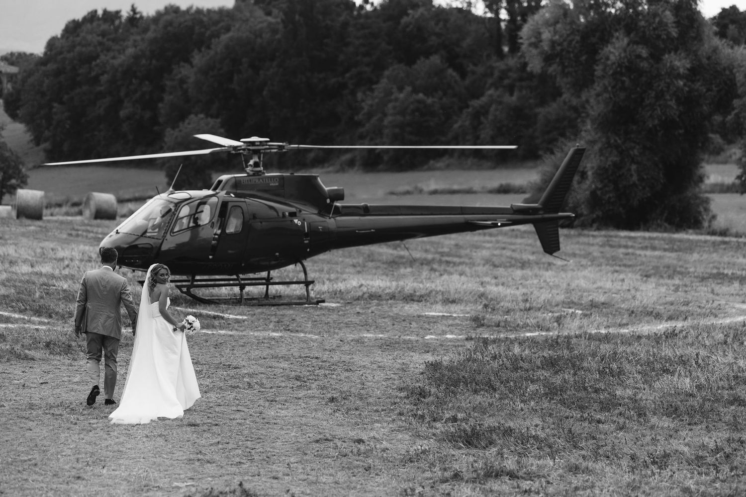 A military helicopter rests in a grassy field at either sunrise or sunset while a person in white walks nearby.