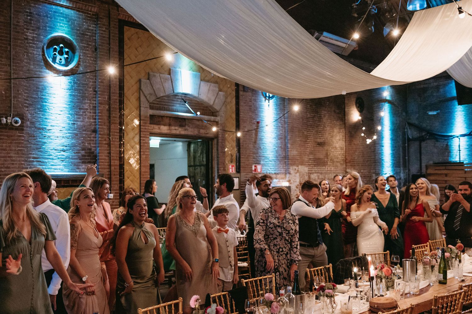 Wedding guests stand and celebrate during reception in venue with dramatic blue lighting and exposed brick walls.