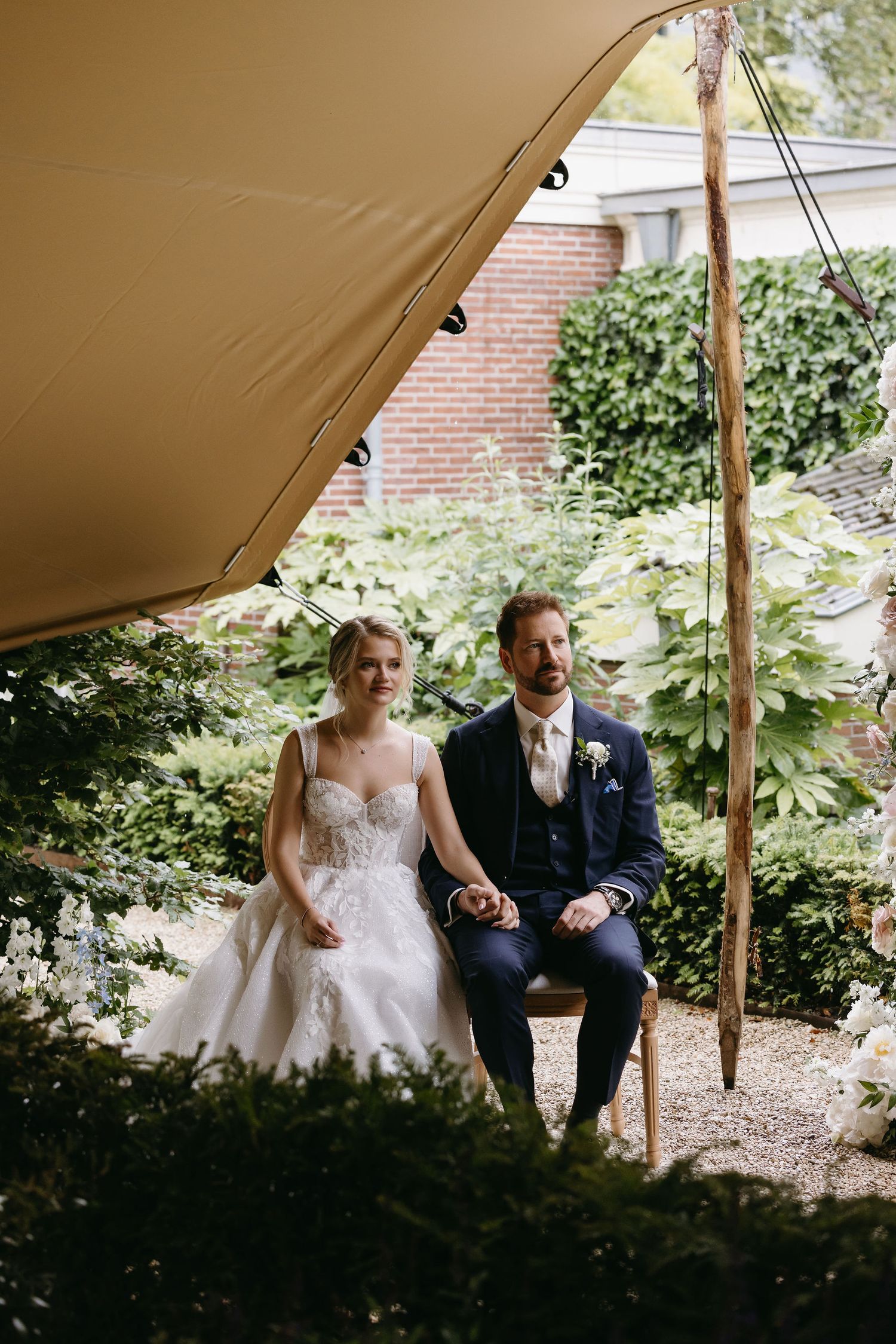 Couple seated under a beige outdoor tent canopy in a garden setting.
