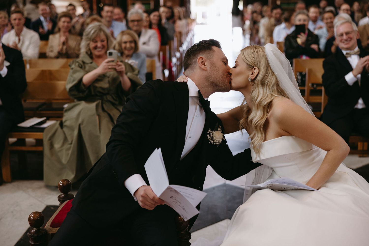 Romantic wedding moment captured in church with guests applauding in background.