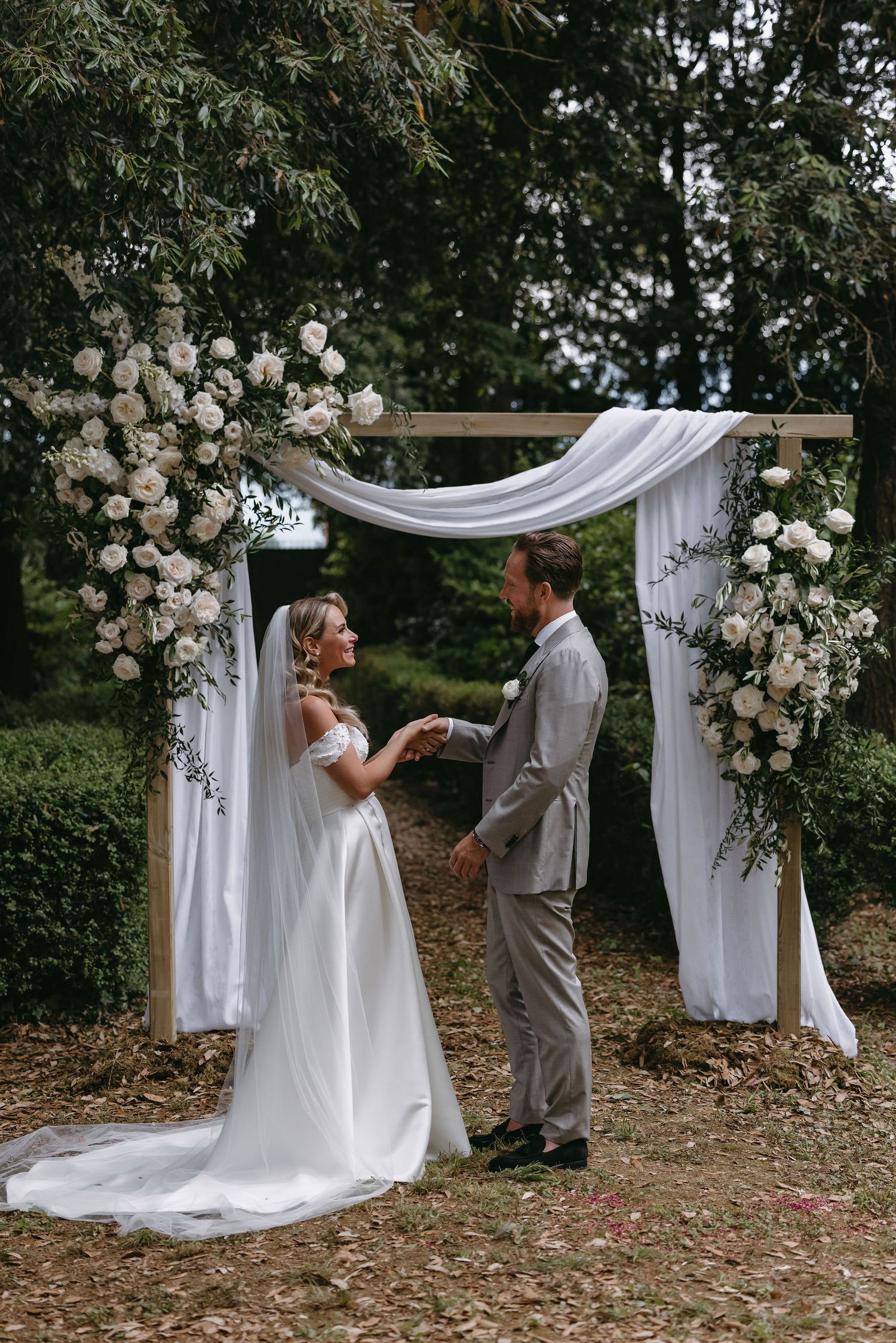Couple exchanges vows under floral arch with white roses and draped fabric.