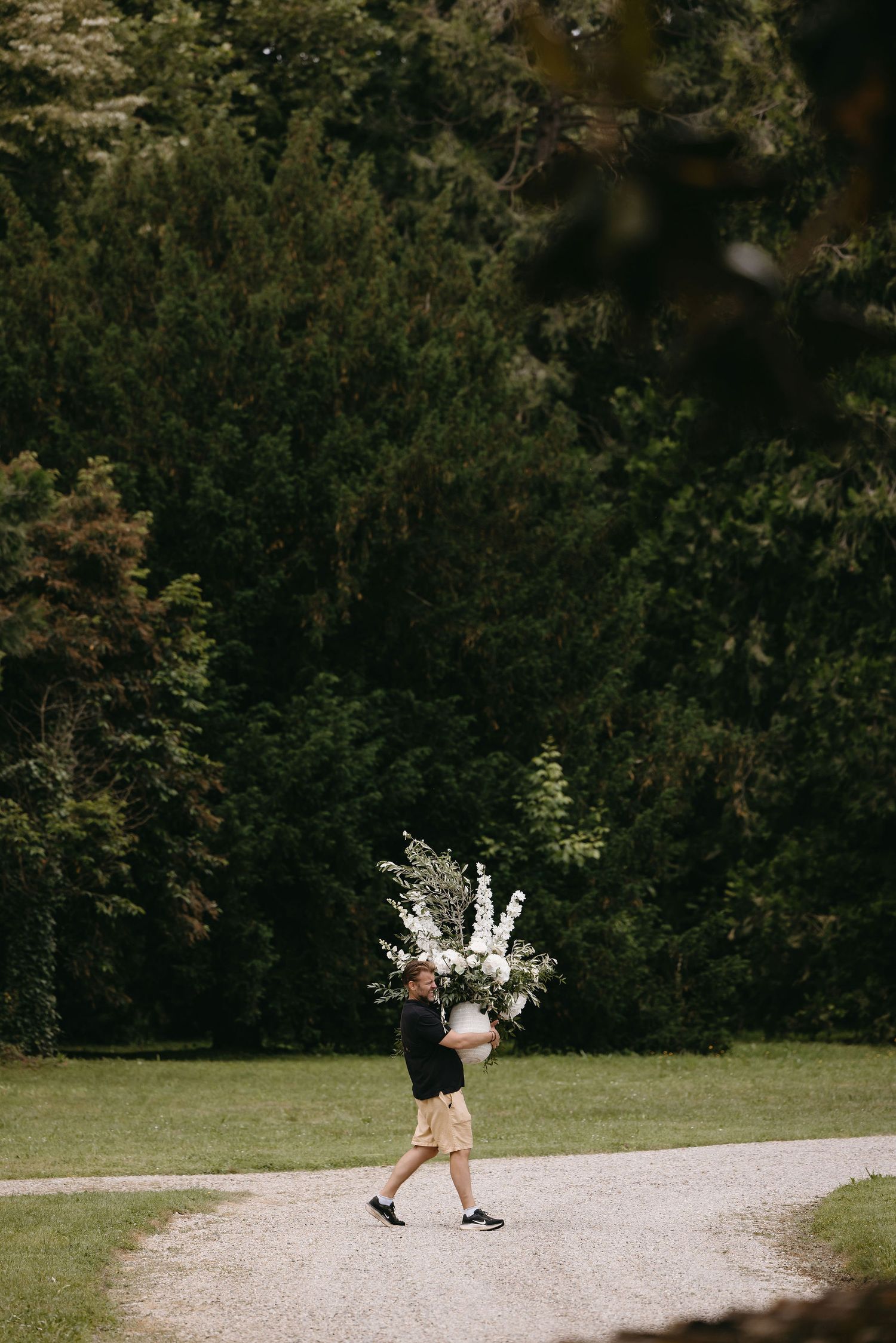 Figure walking on a path carrying a large bouquet of white flowers against a dark green forest background.