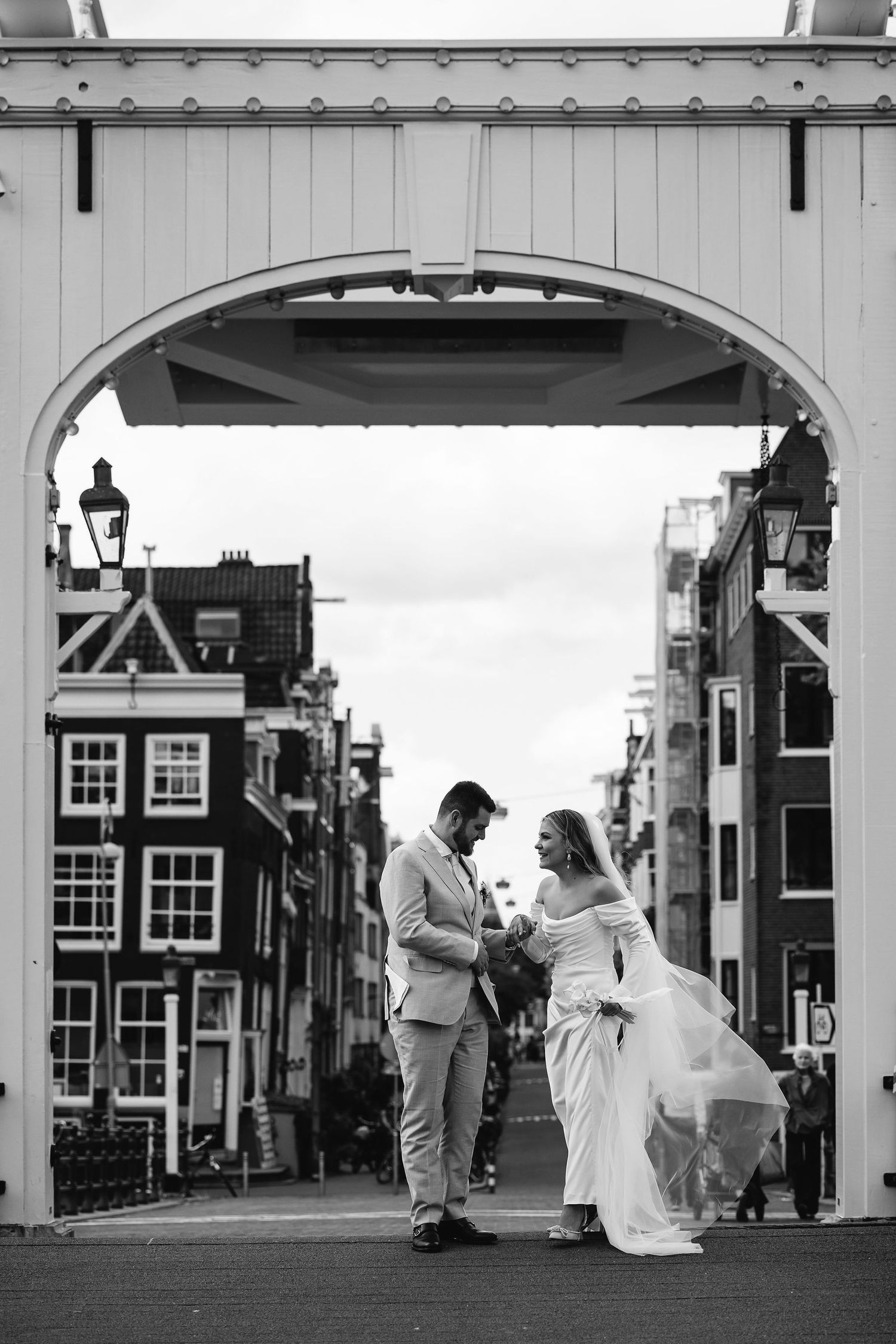 A romantic black and white wedding photo of a couple under an arched bridge in Amsterdam with classic architecture.