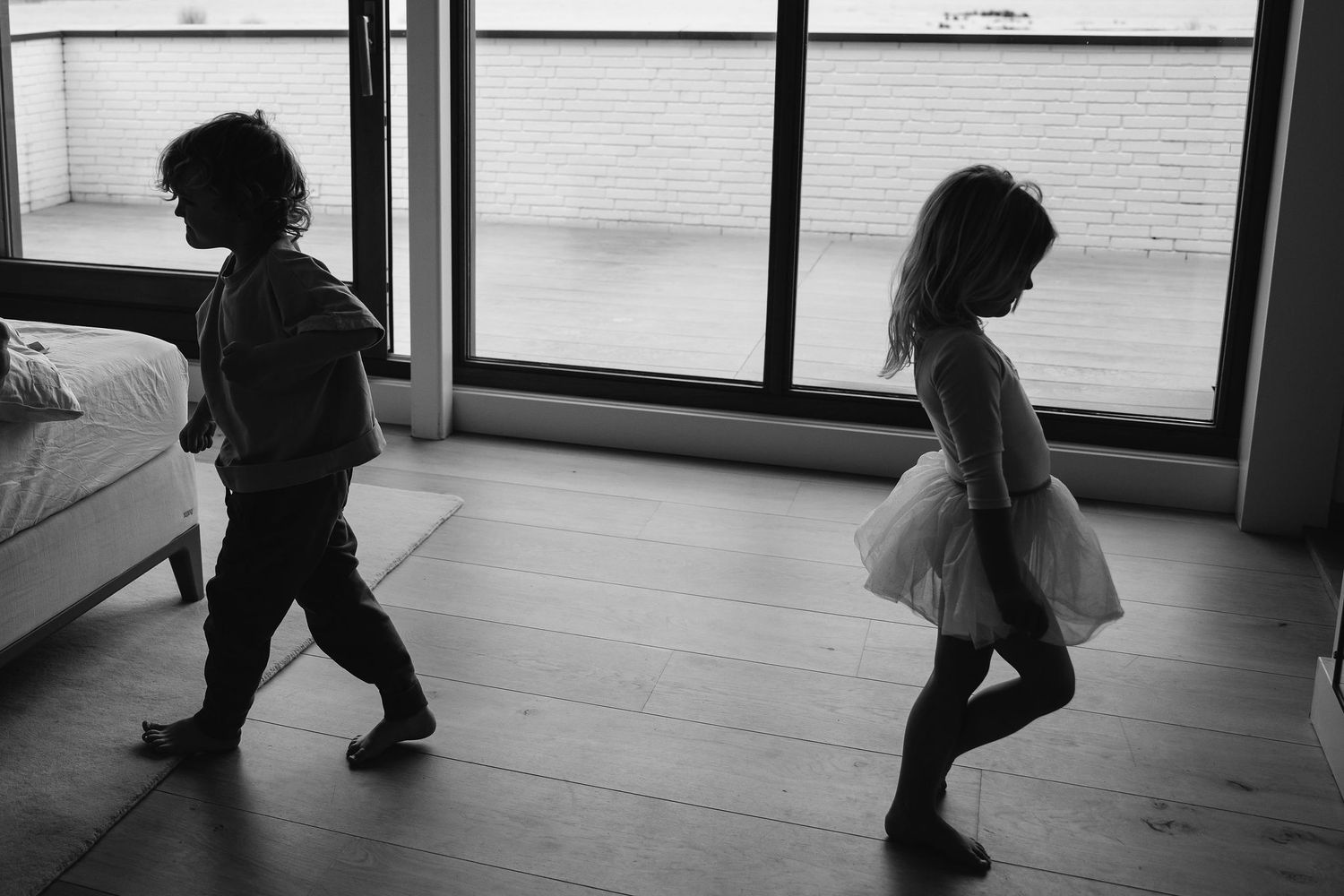 Two children dancing and playing in a room with floor-to-ceiling windows.
