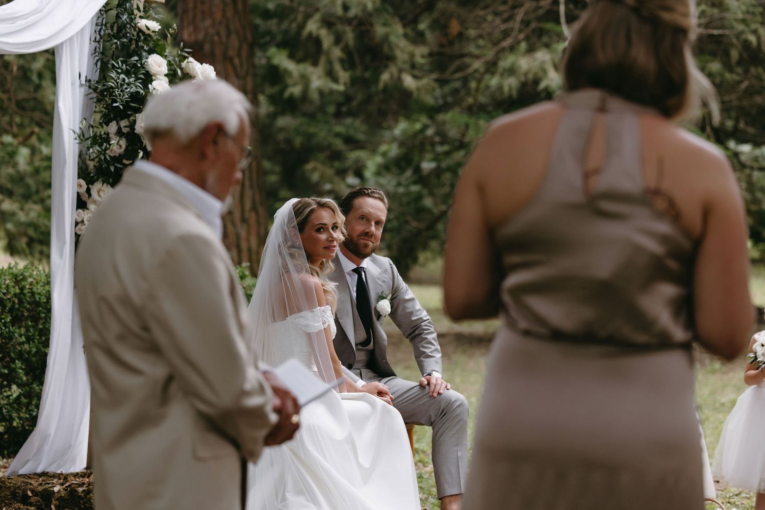 Outdoor wedding ceremony under white draped arch with natural forest backdrop.
