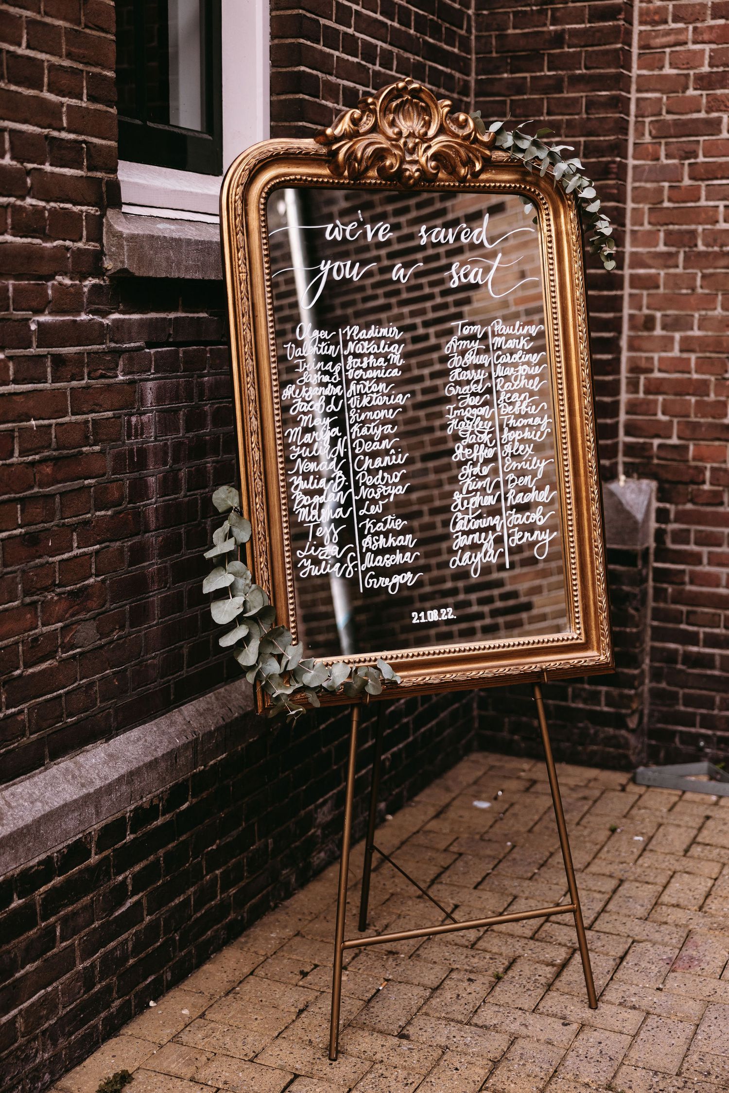 Elegant gold-framed mirror displays wedding seating chart against brick wall.