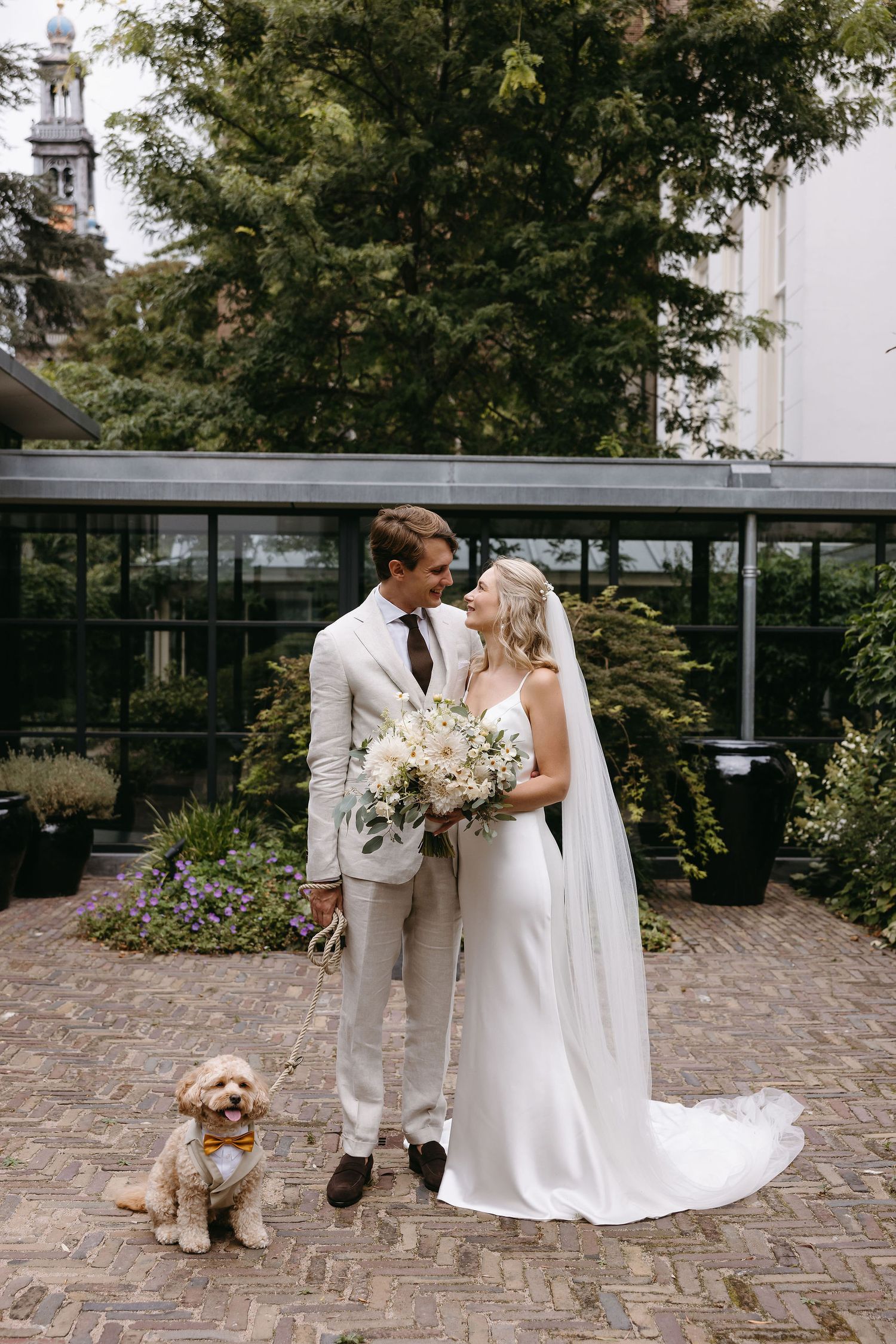 Elegant couple in white attire pose for wedding photos with their faithful dog by their side.