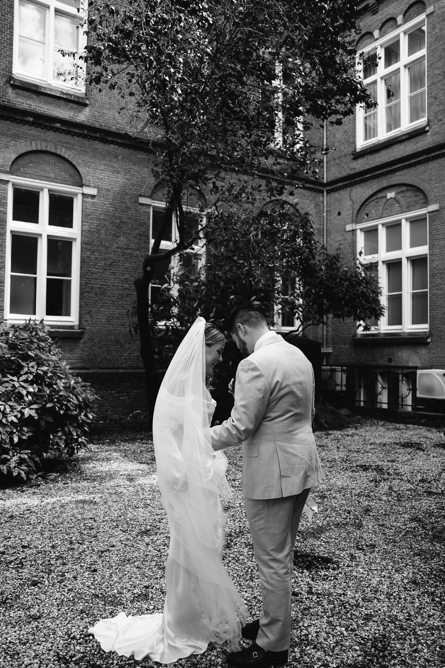 Black and white wedding portrait in courtyard with brick building and trees.