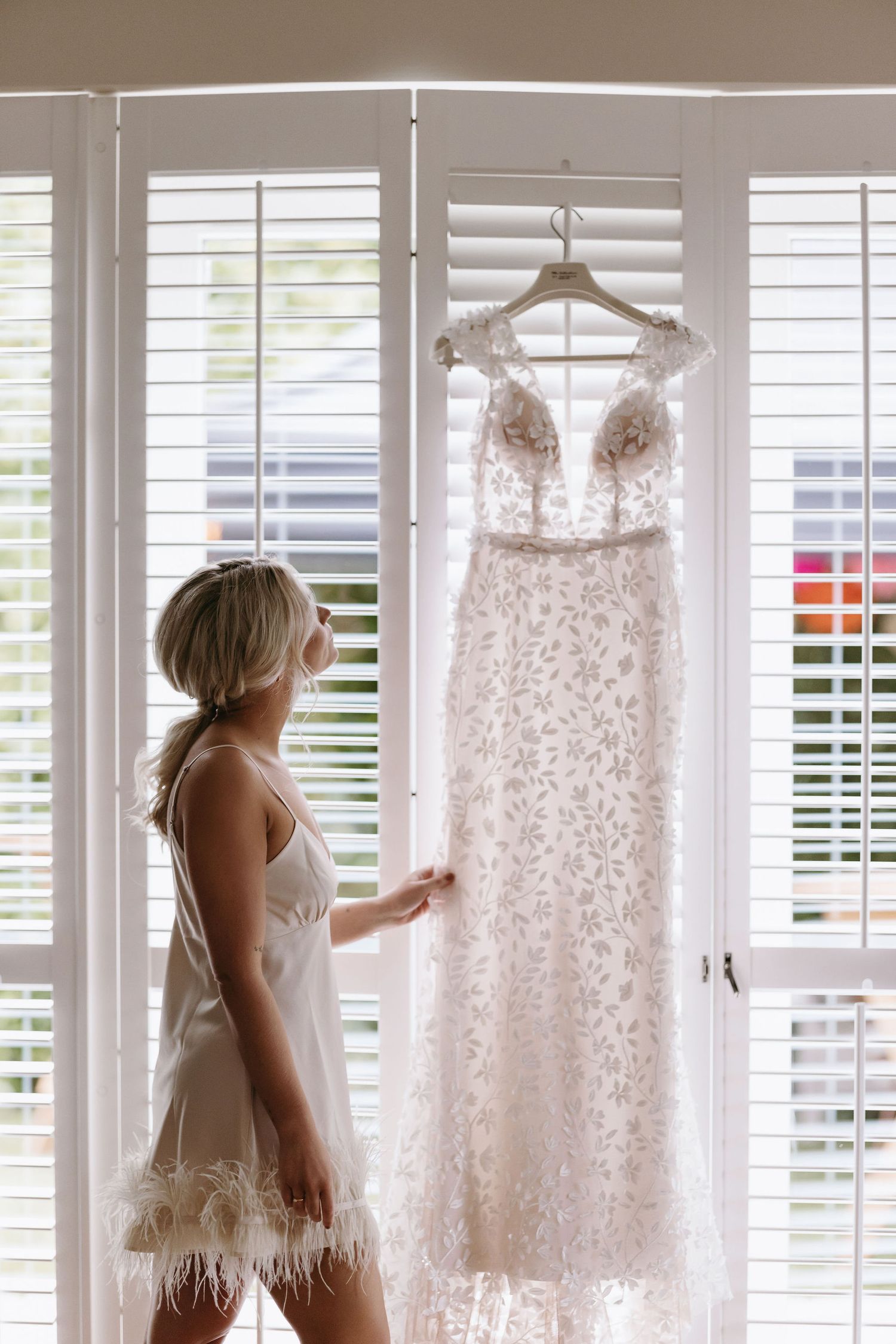 Wedding dresses hanging on white shuttered windows in bright interior space.