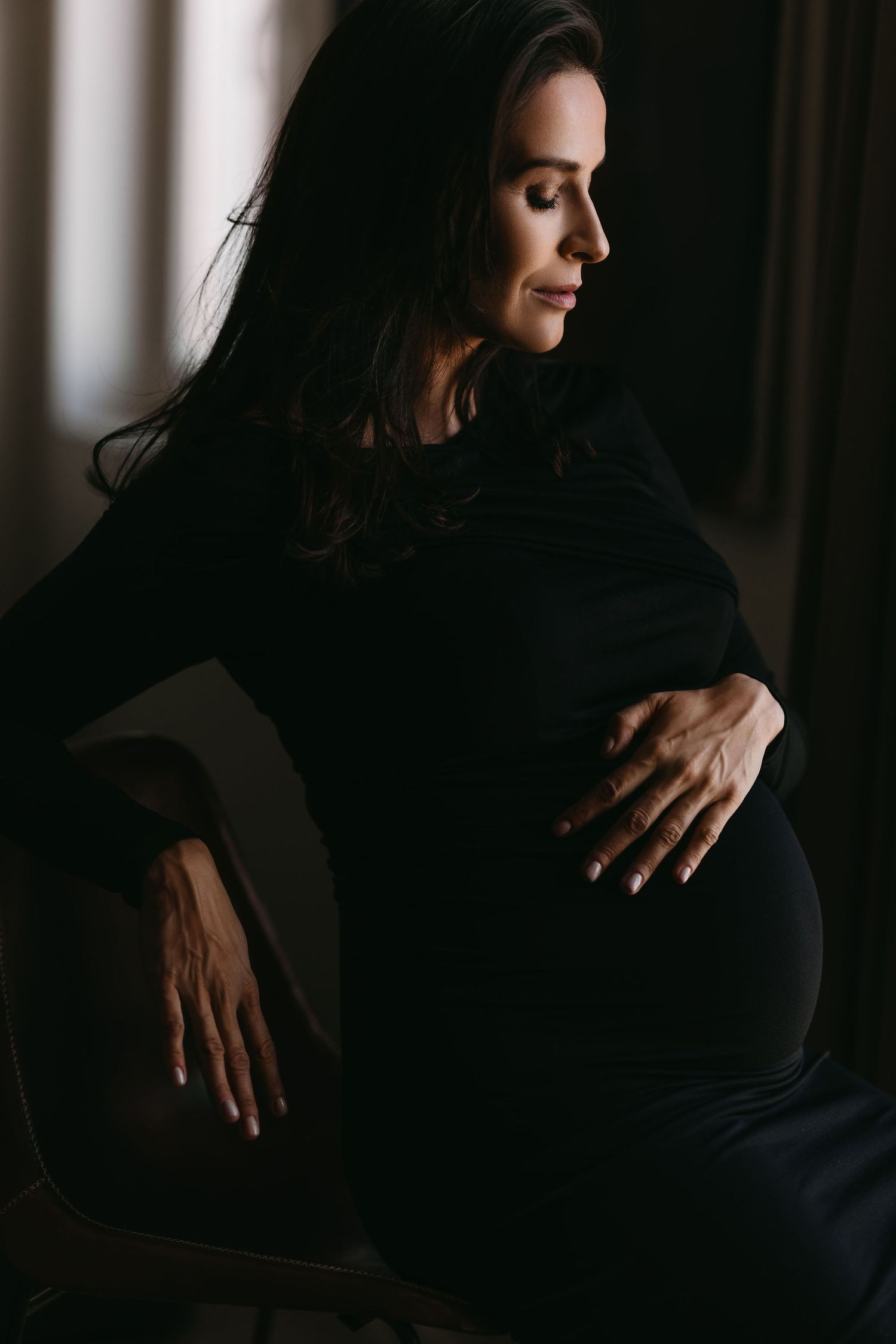 A moody dark maternity portrait of a pregnant woman in silhouette holding her belly with gentle hands.