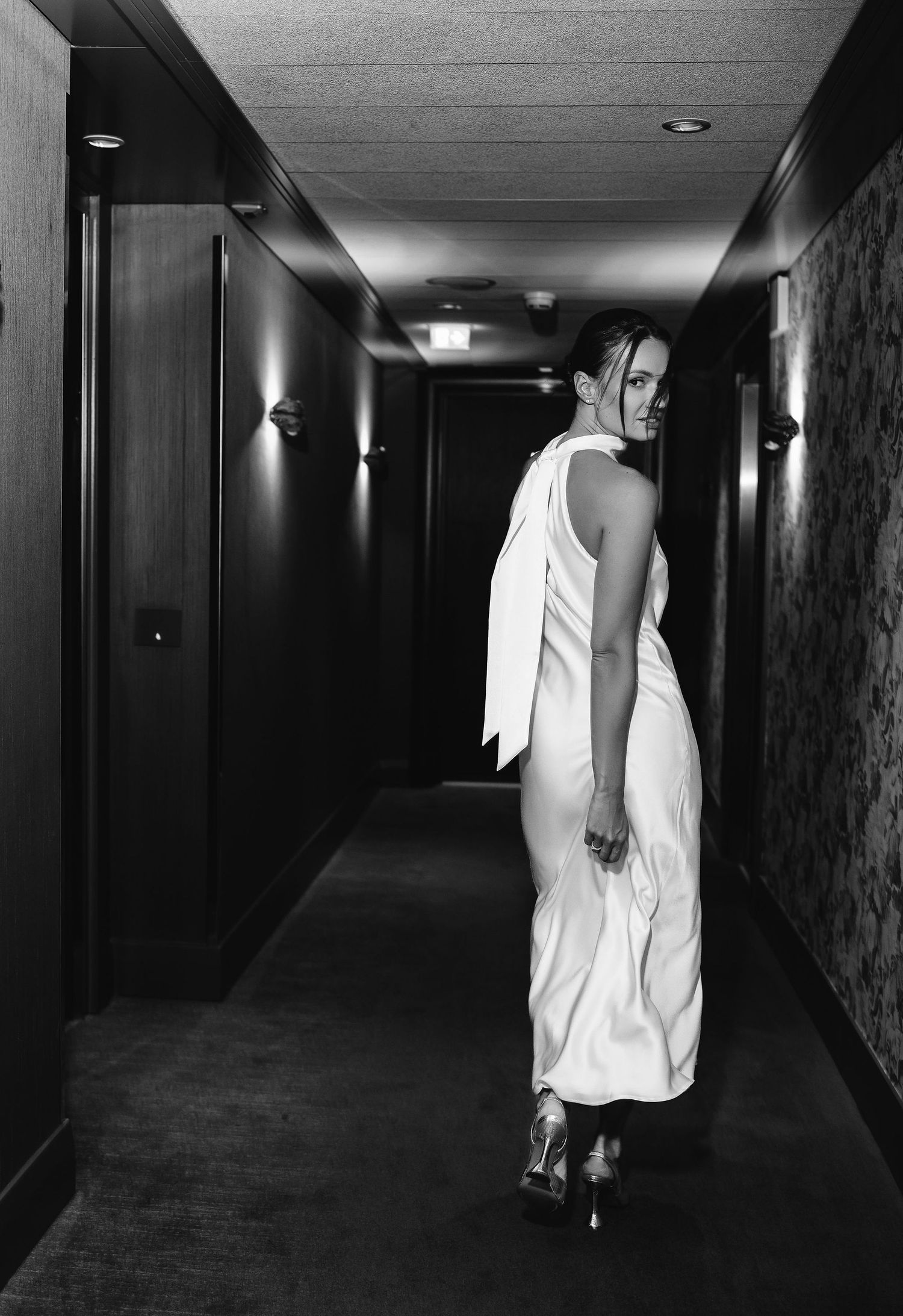 A black and white photo shows a figure in a flowing white dress walking down a dimly lit hotel hallway.