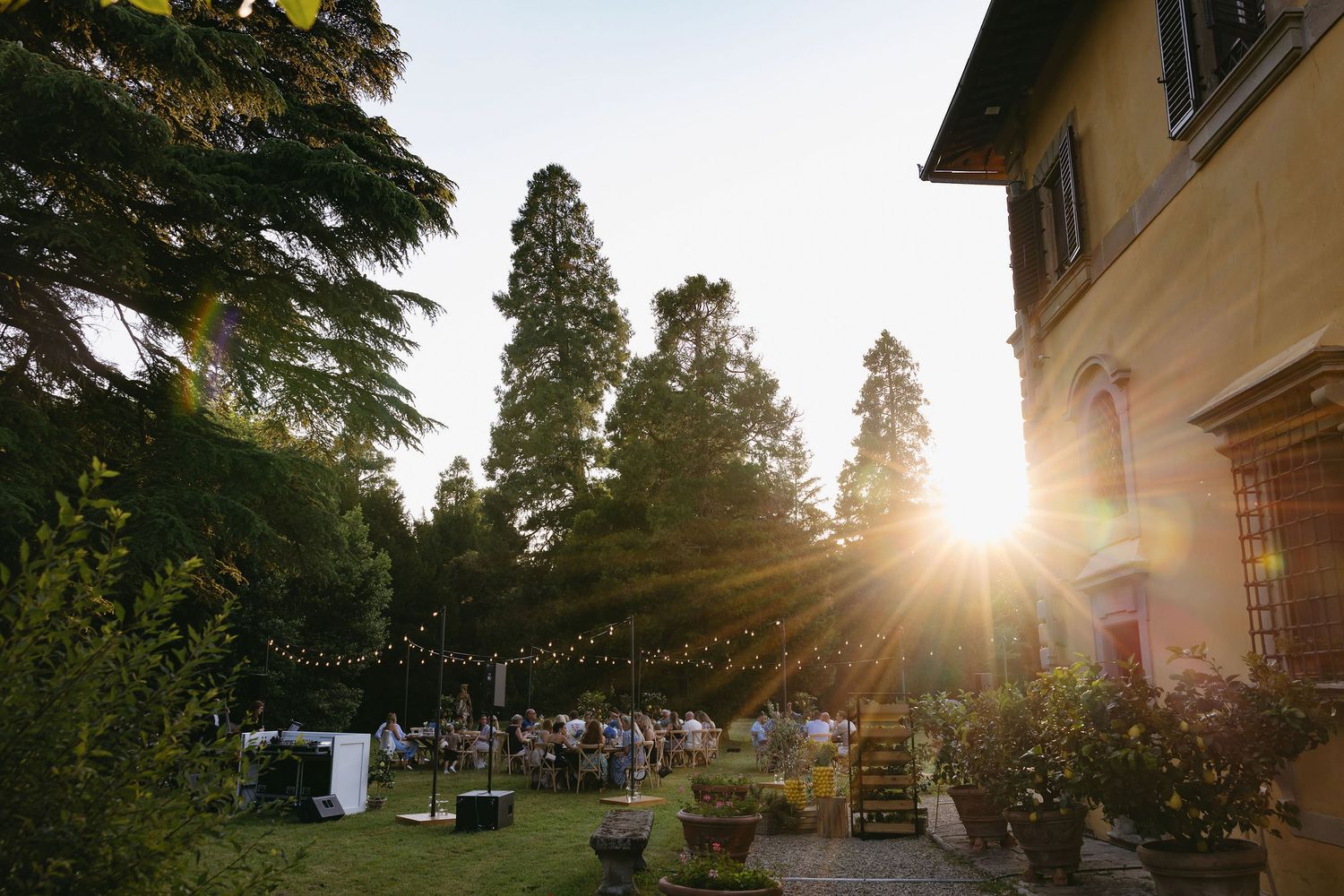 A sunset view of an outdoor wedding reception setup with tall trees and a villa in the background.