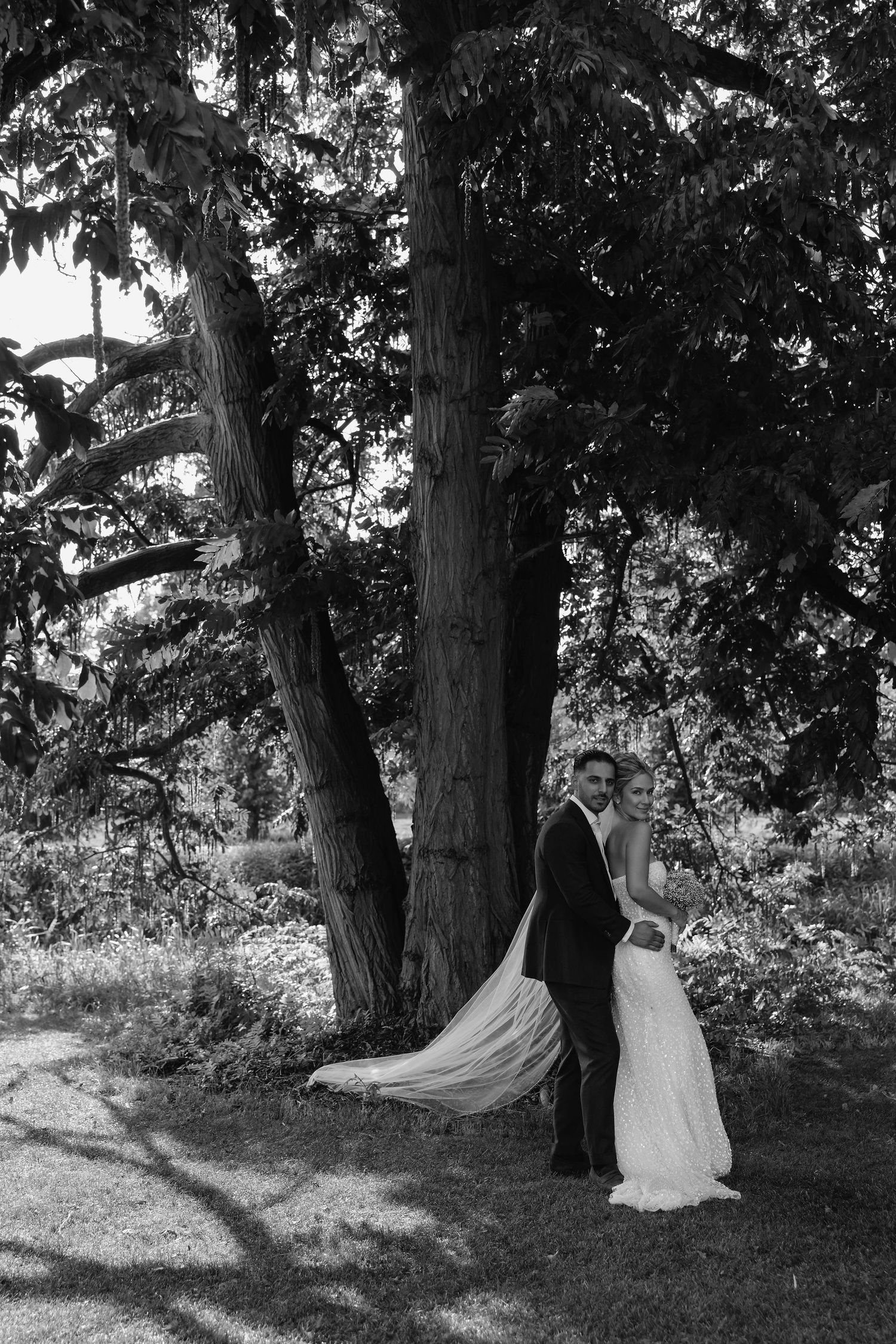 A black and white wedding photo showing a couple standing by a large tree in a natural outdoor setting.