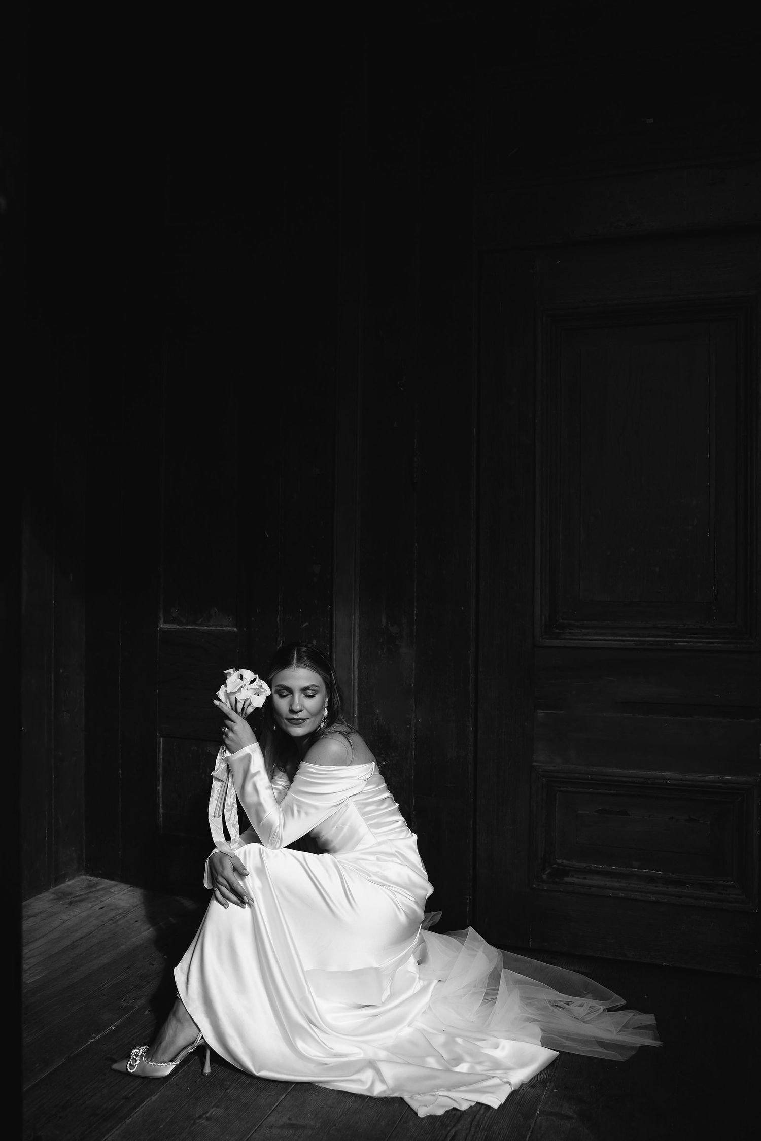 A bride in an elegant white off-shoulder gown sits on the floor holding a bouquet in a dramatic black and white portrait.