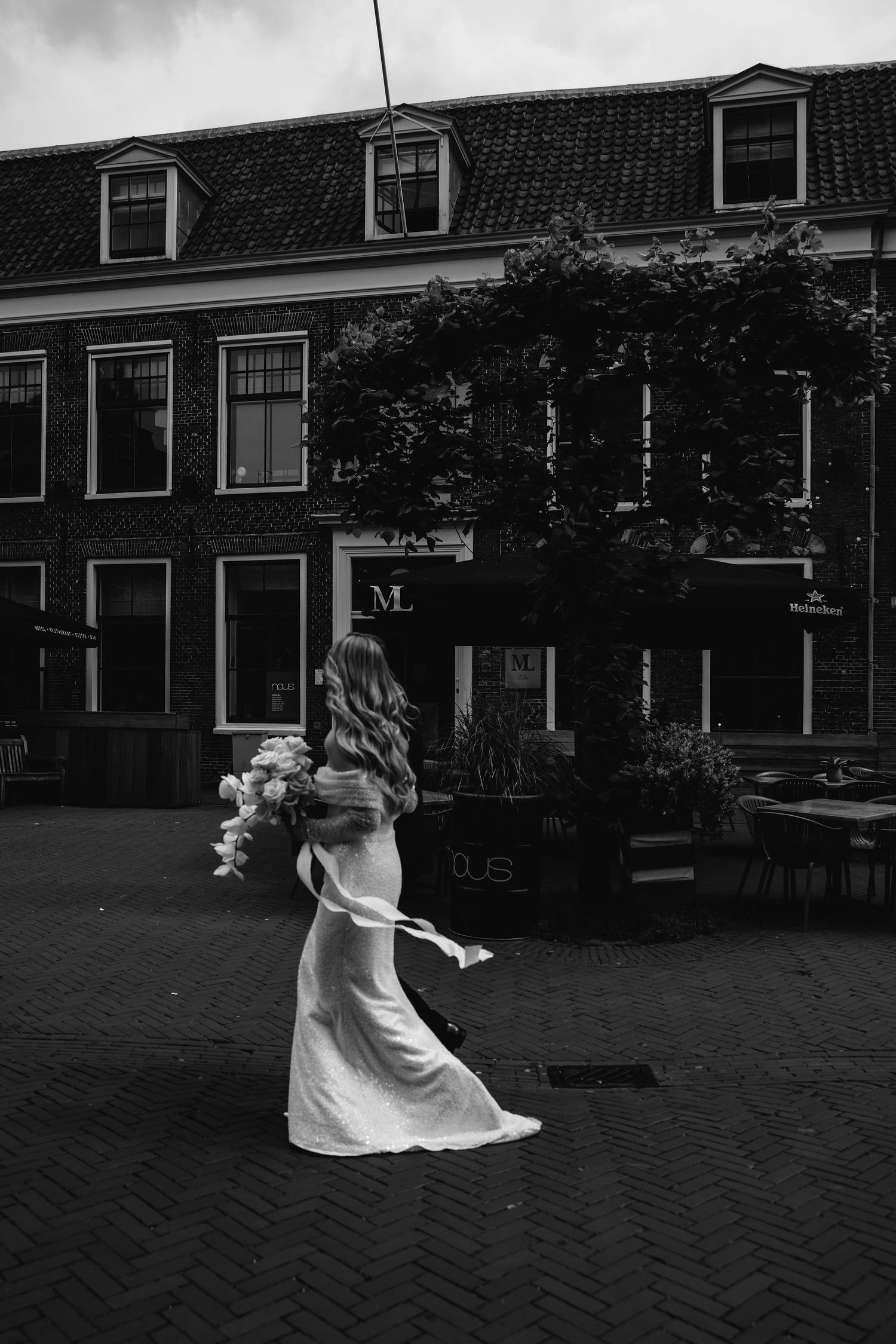 A figure in a flowing white dress dances on cobblestone streets beside historic Dutch-style buildings in black and white.