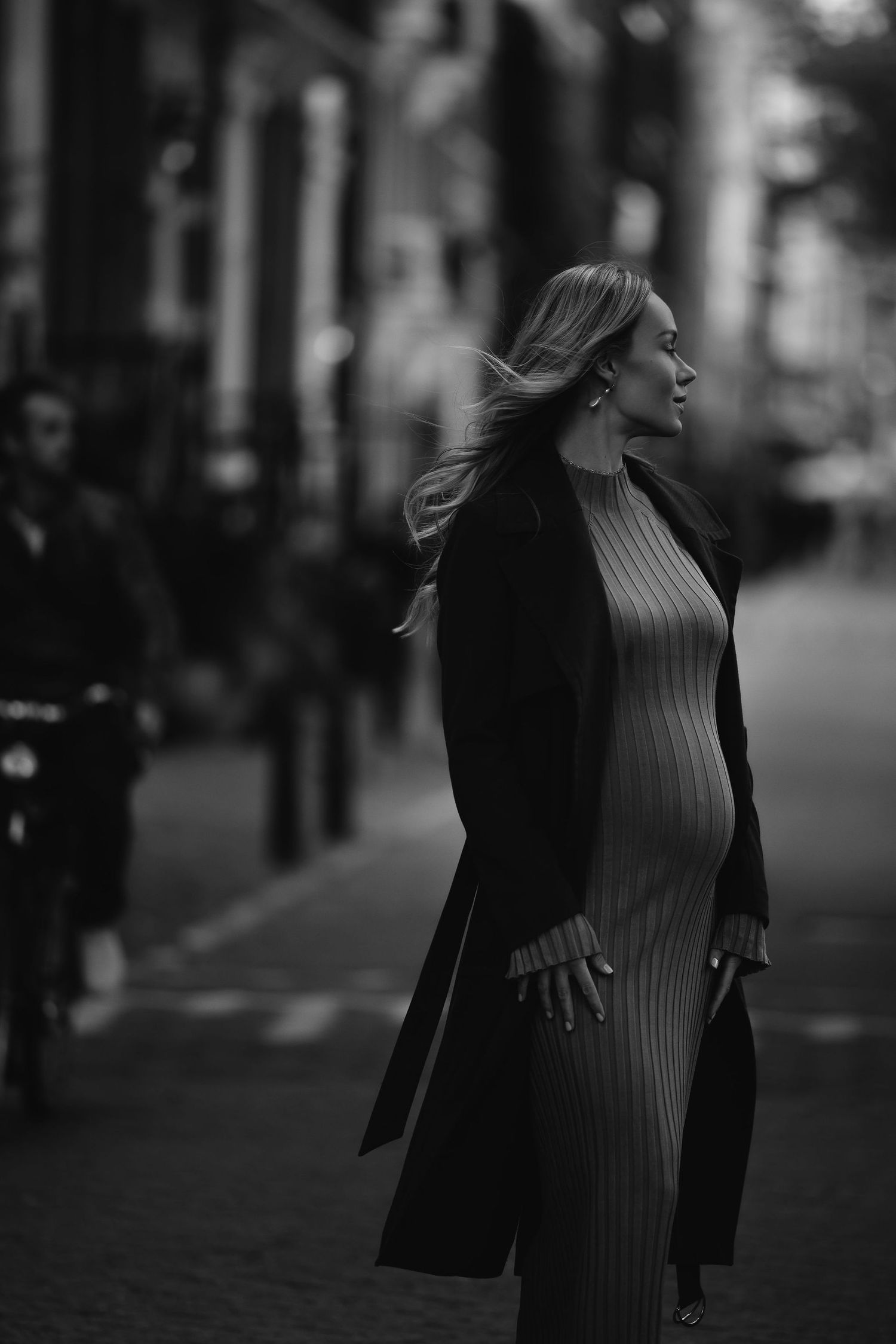 Black and white street photo of pregnant woman in ribbed dress and long coat.