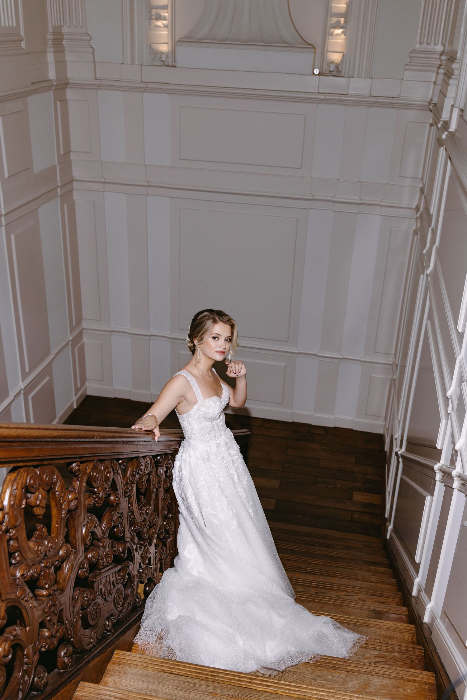 Bride in a flowing white lace gown poses elegantly on ornate wooden stairs.