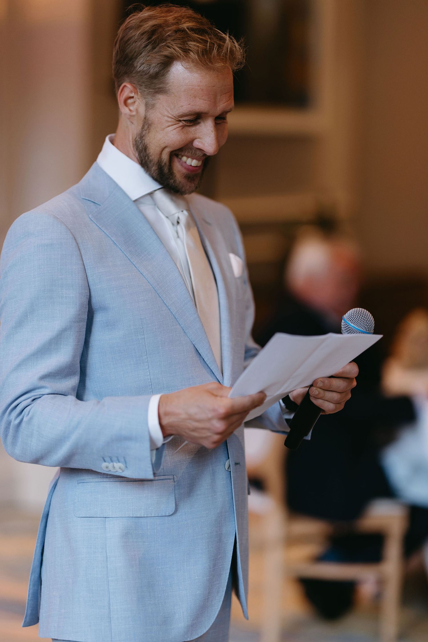 A man in a light blue suit reads from papers during a wedding speech.