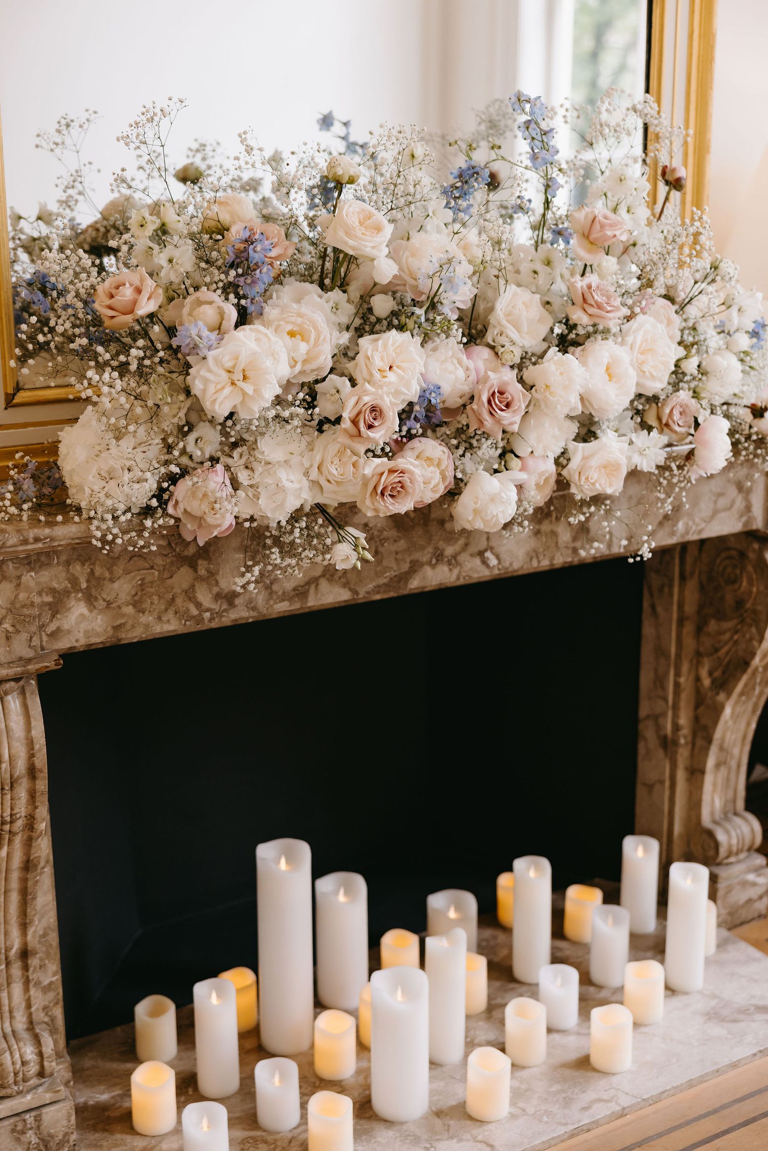Romantic floral arrangement of white roses and baby's breath above glowing candles on a marble fireplace.