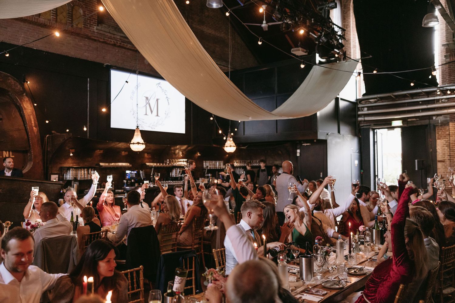 Wedding reception hall filled with guests and decorated with hanging fabric and lights.