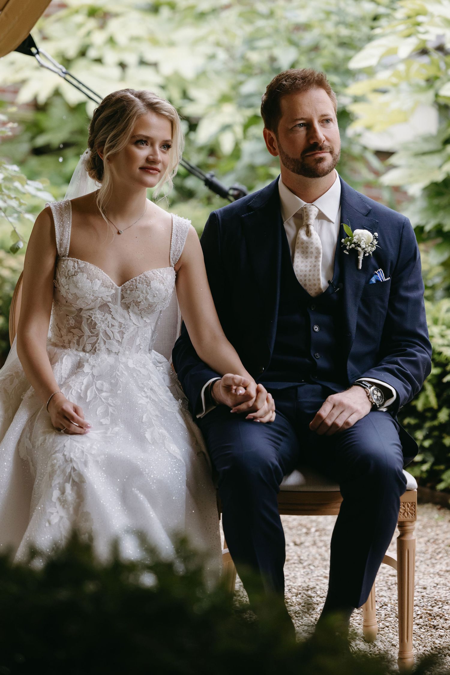 Newlyweds sitting together outdoors, with bride in lace dress and groom in navy suit.