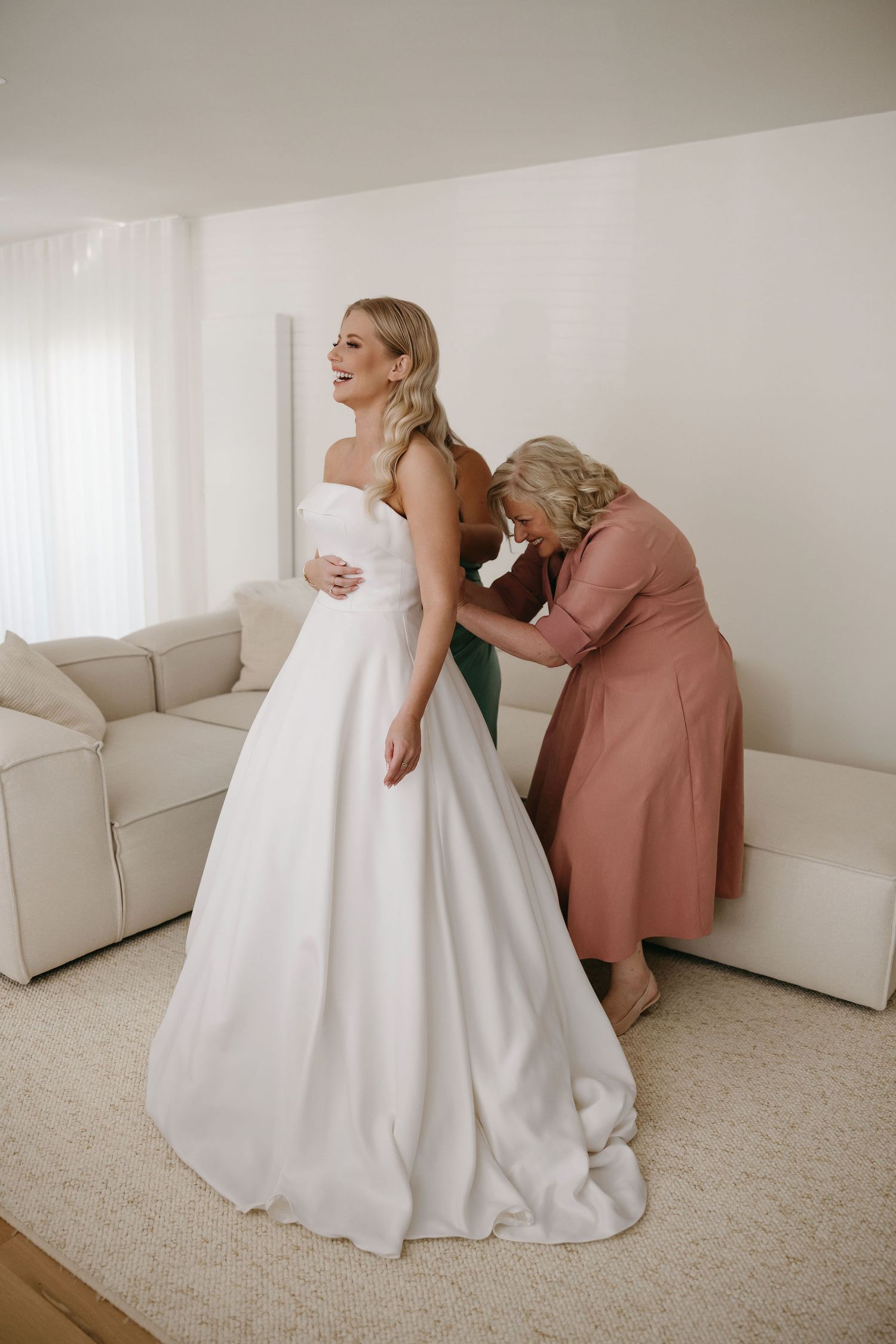 A bride in a classic white wedding gown gets her dress adjusted in a light-filled room with a beige couch.