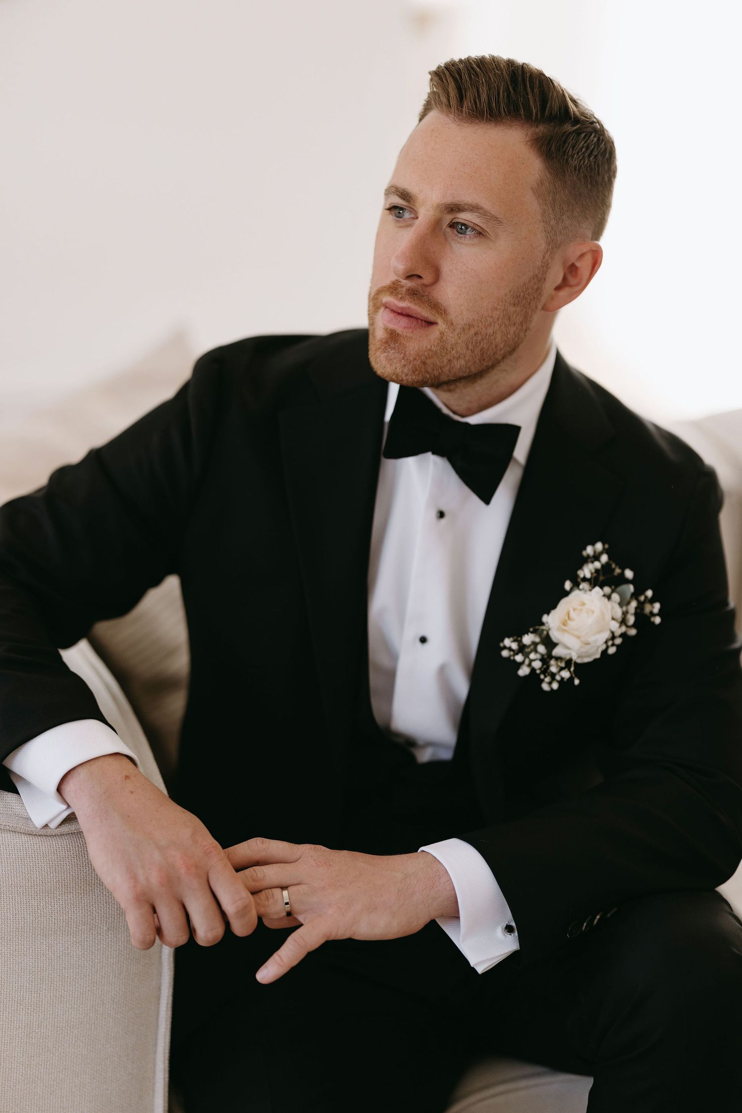 Person in black tuxedo and white boutonniere sits elegantly on light colored furniture.