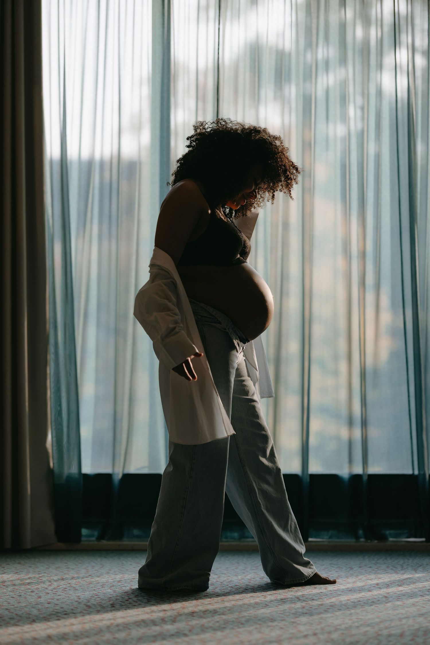Side profile of pregnant figure in casual wear standing by sheer curtains.