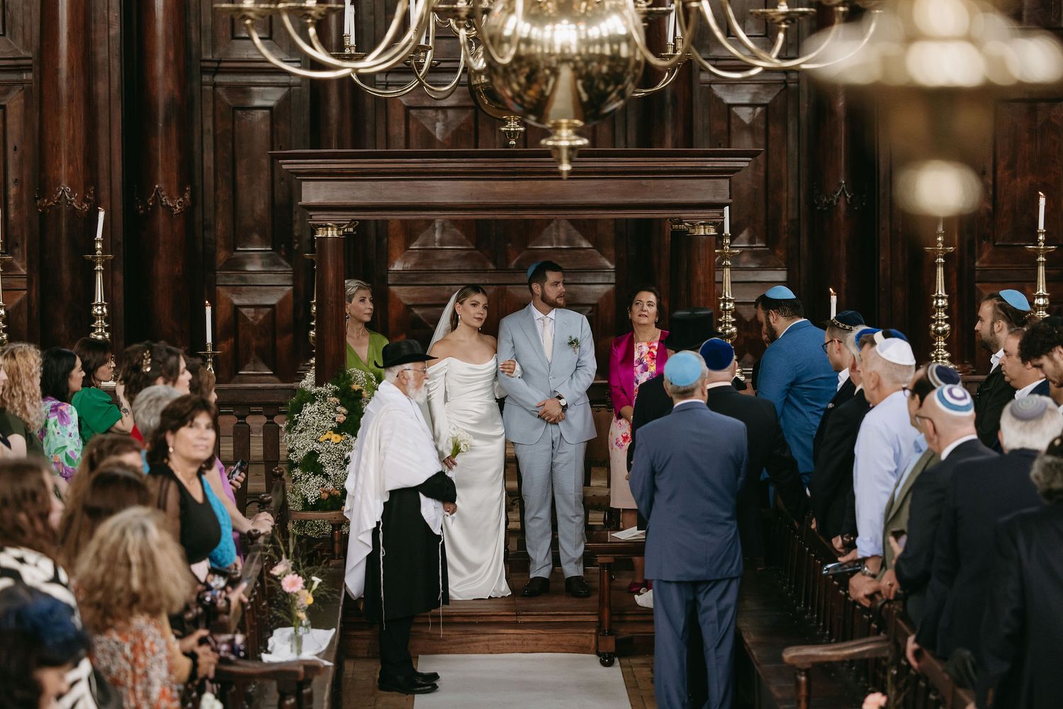 Wedding ceremony in ornate wood-paneled room with chandelier and gathered guests.