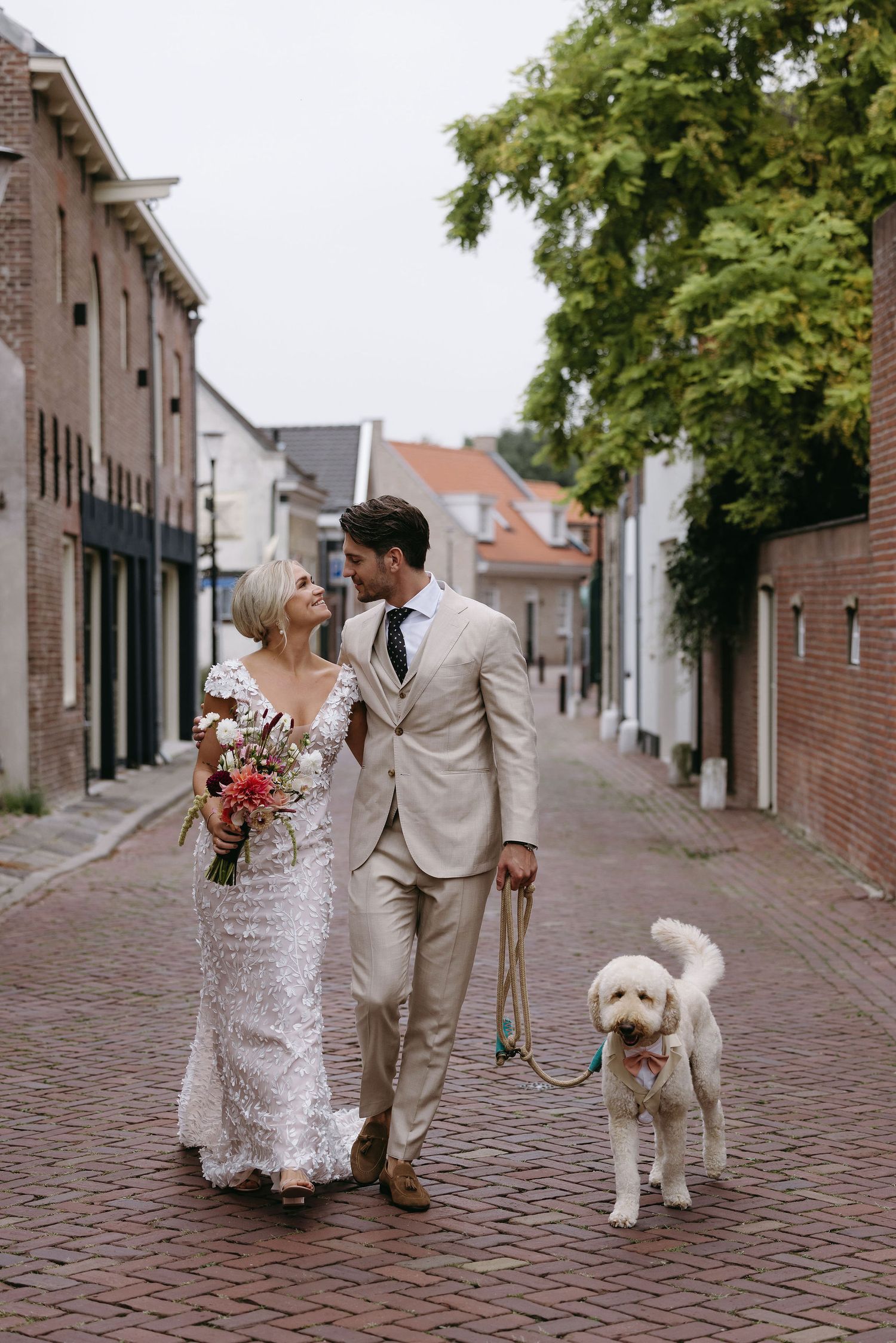 Couple walking down cobblestone street with white dog during wedding photoshoot.