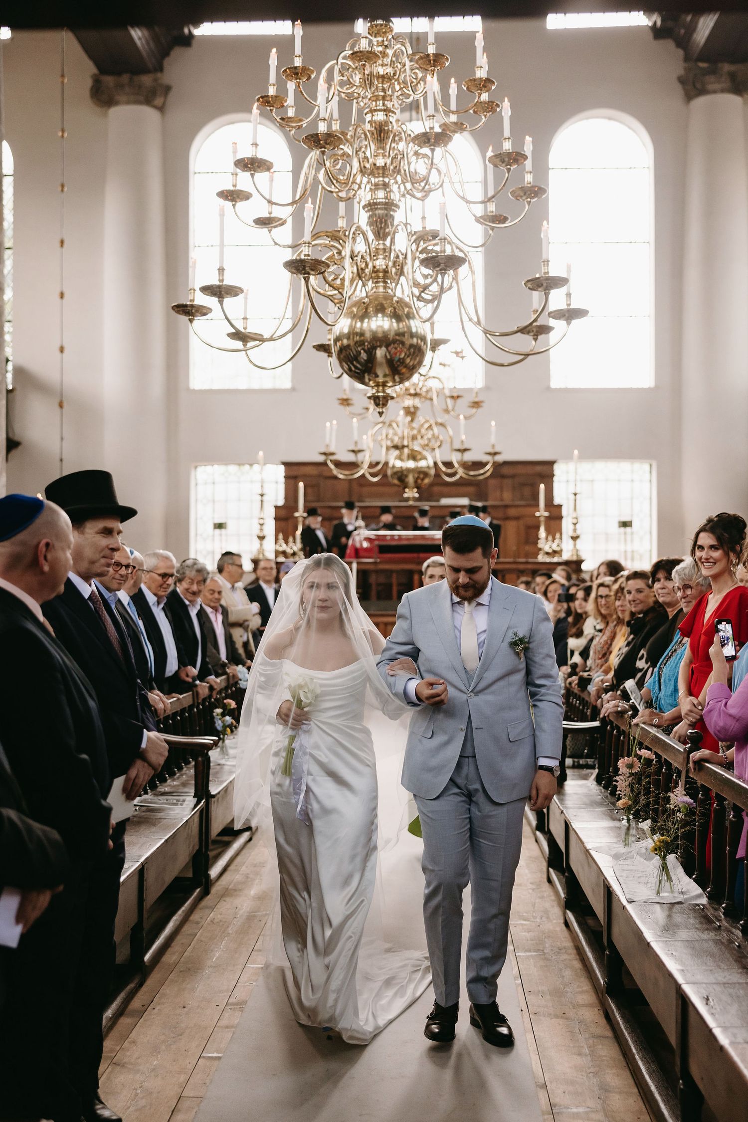 Wedding ceremony in a bright church interior with elegant chandeliers and wooden pews filled with guests.