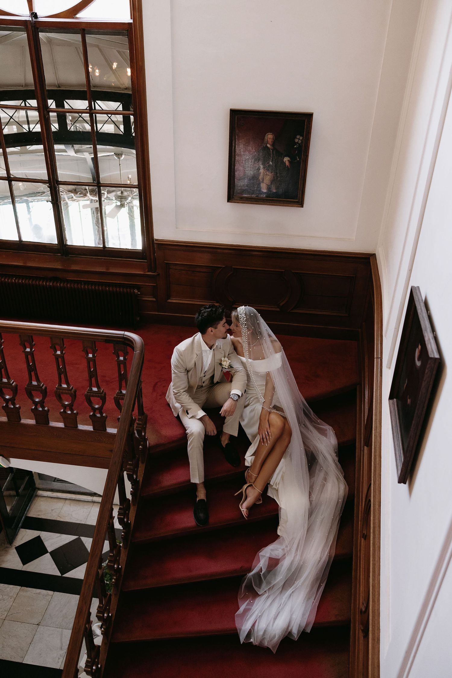 A couple sits intimately on elegant red-carpeted stairs in a historic building with wood paneling and vintage decor.