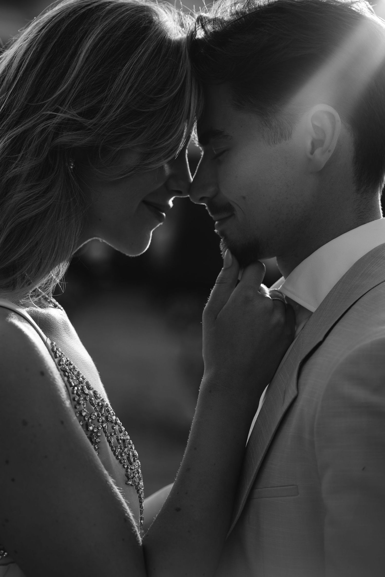 A romantic black and white close-up photo of a couple's intimate moment in formal attire with sunlight behind them.