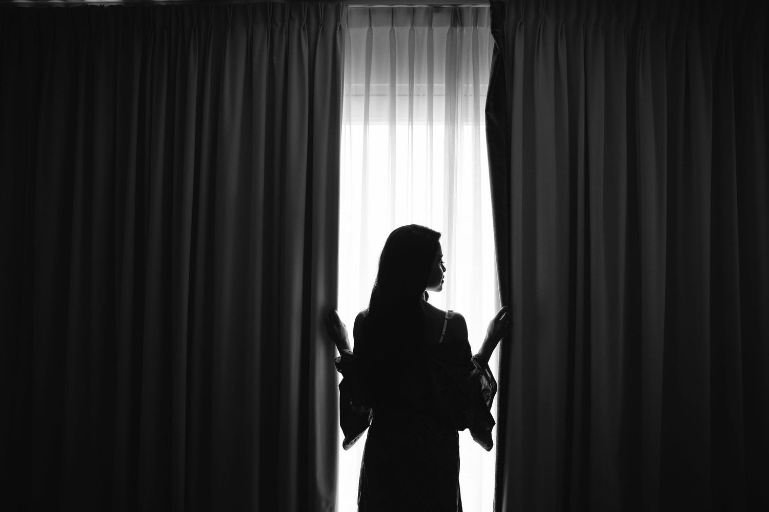 A silhouetted figure stands between curtains against bright backlight in dramatic black and white composition.
