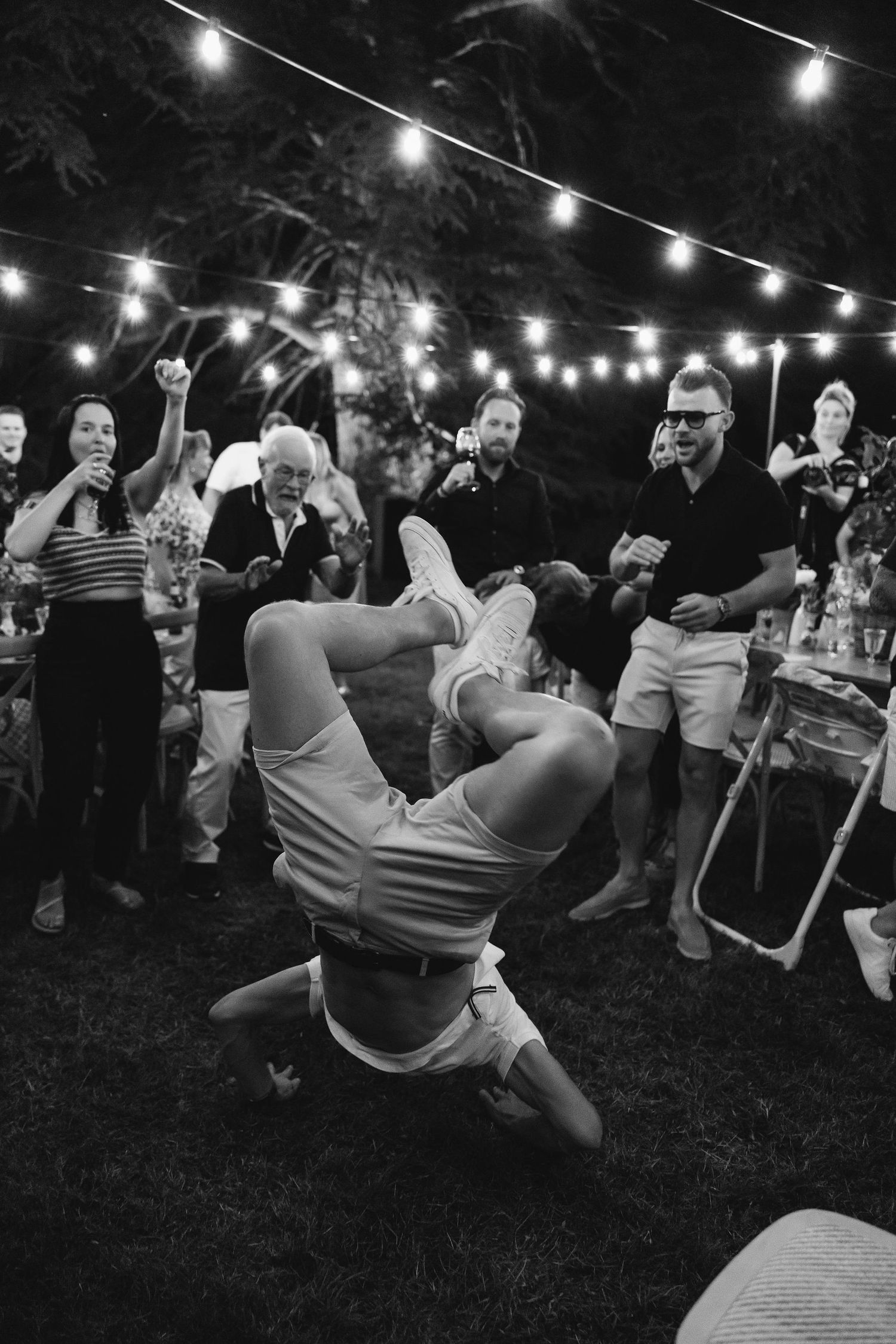 Black and white photo of wedding guests enjoying outdoor party under string lights.