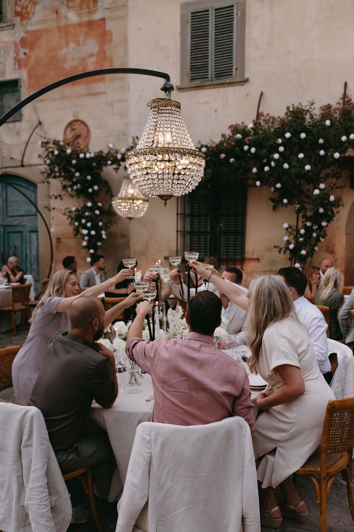 An elegant outdoor dinner party with guests toasting under a crystal chandelier against a rustic Italian villa backdrop.