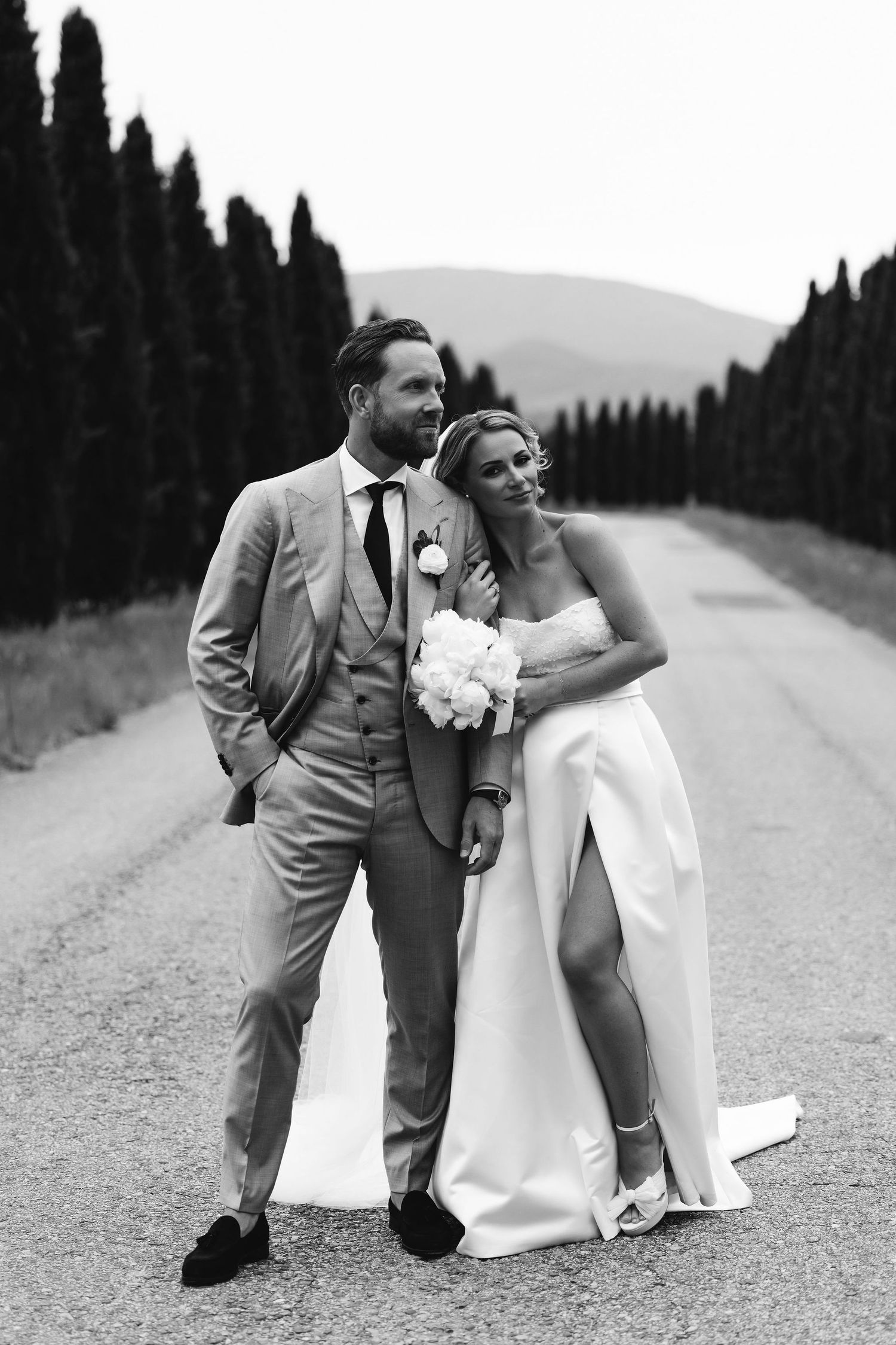Romantic wedding couple poses on a tree-lined path in black and white photography.