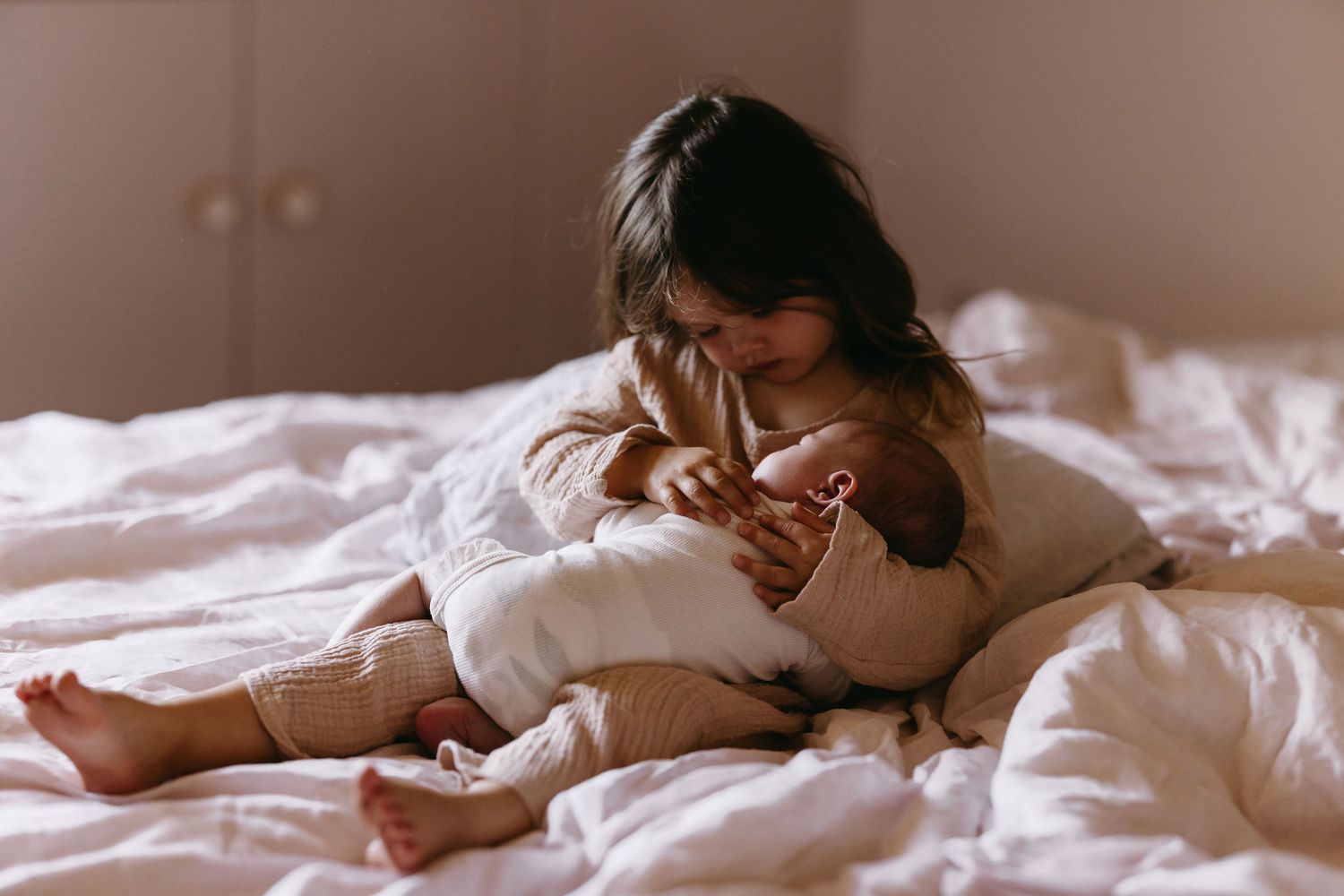 Intimate moment of parent and baby cuddling together on white bedding in soft lighting.