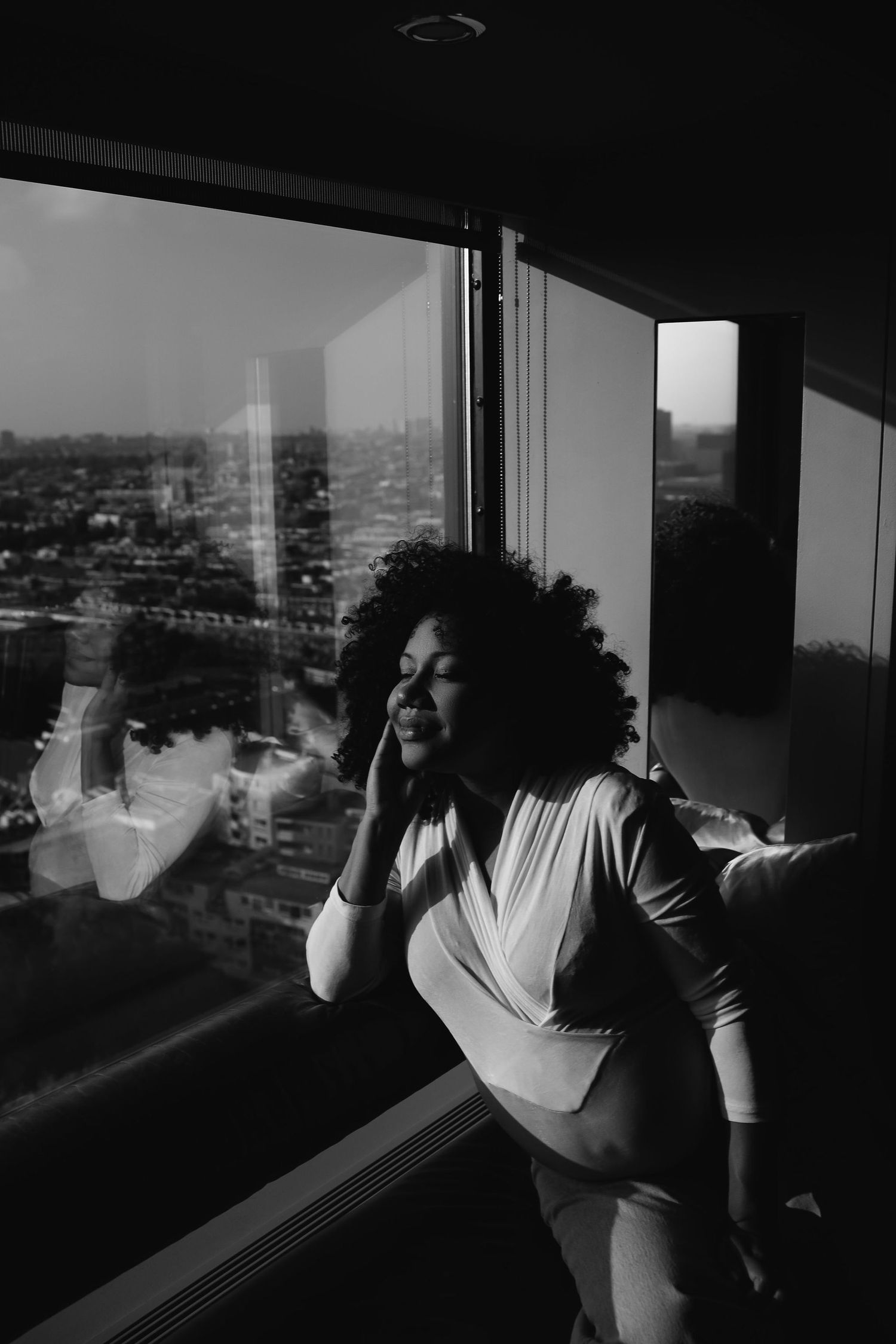 Artistic black and white portrait by a window overlooking an urban landscape.