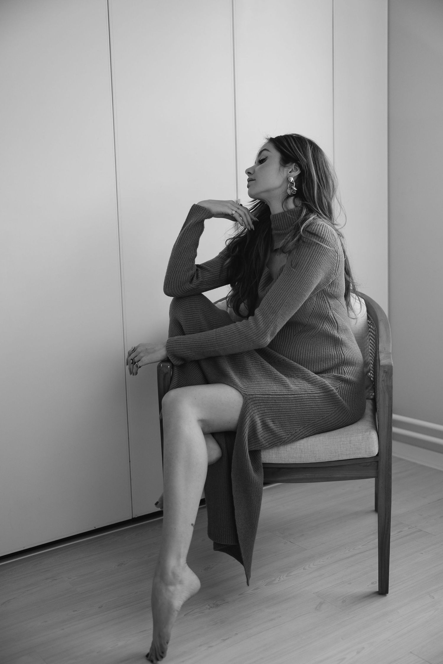 Black and white portrait of woman in sweater dress seated on wooden chair.
