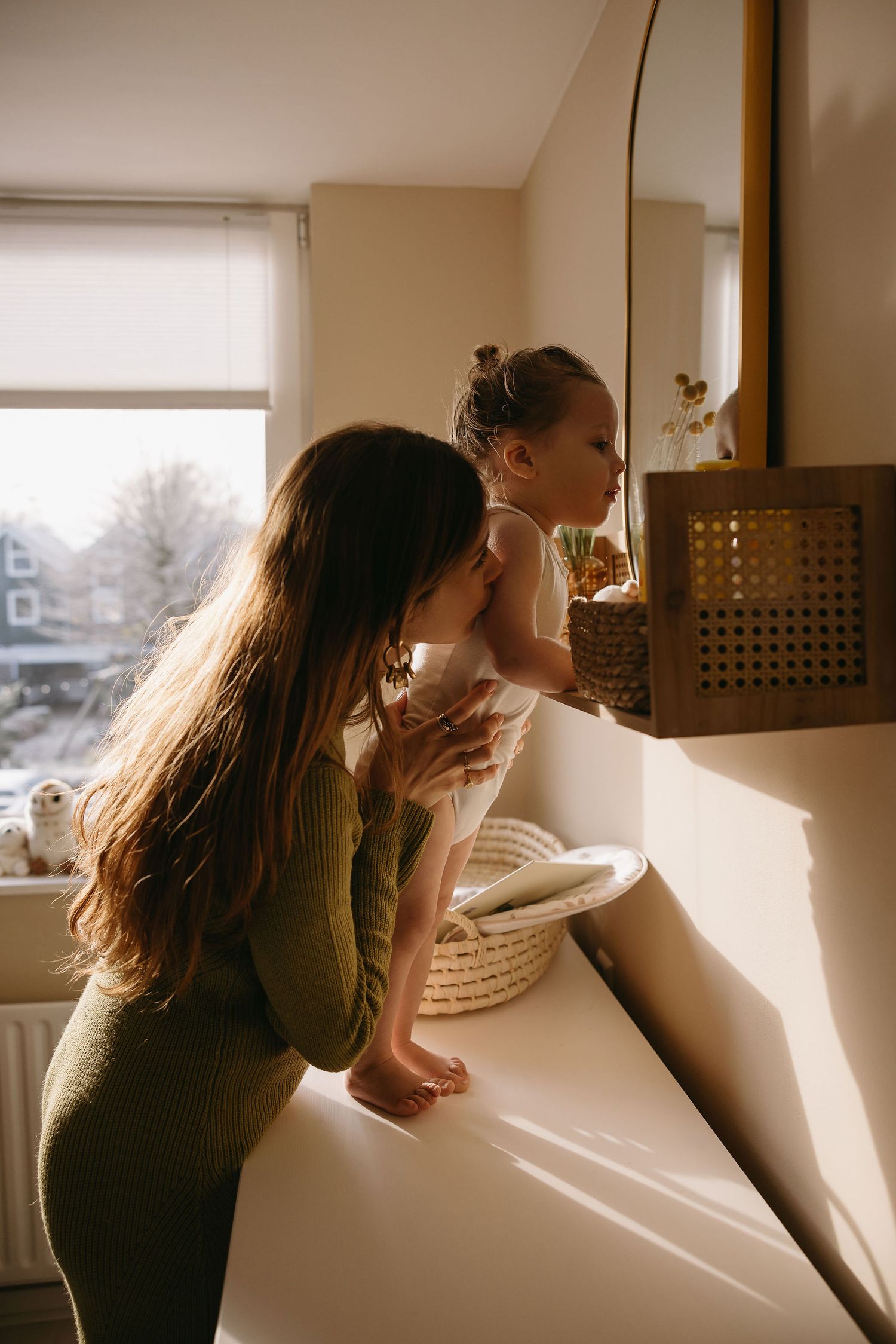 Mother and child look out window together in warm morning light.