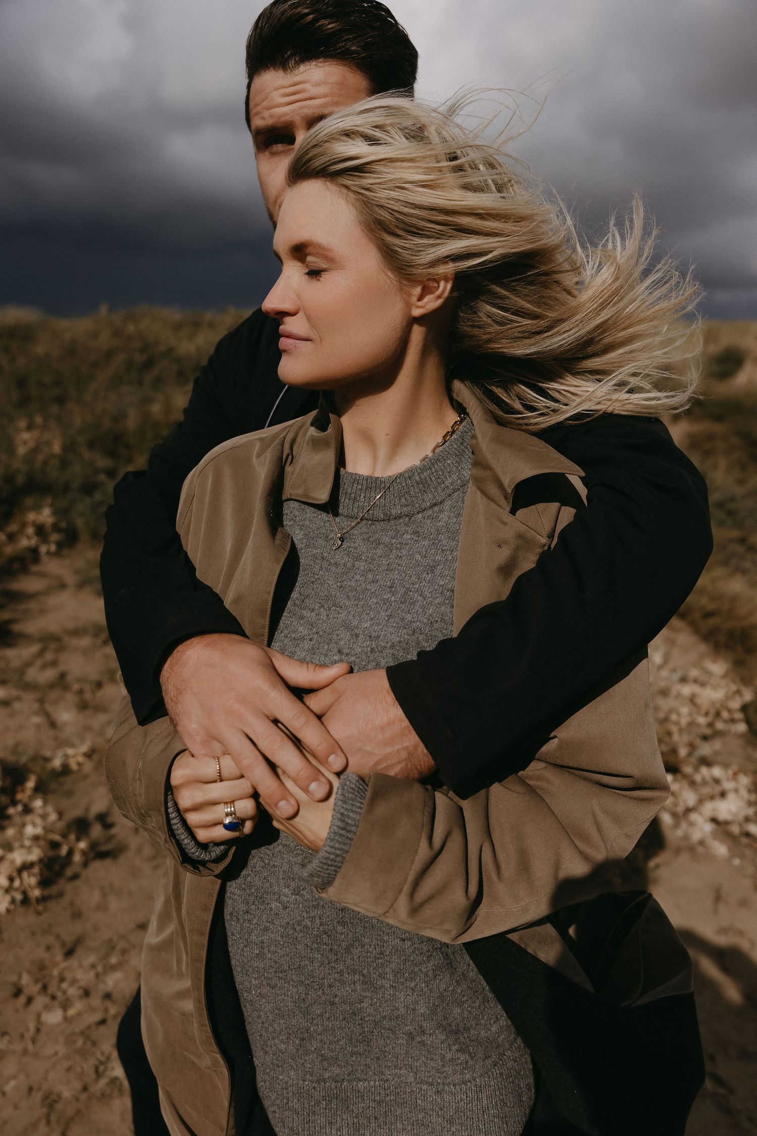 A couple embraces on a windy hilltop with dramatic storm clouds in the background.