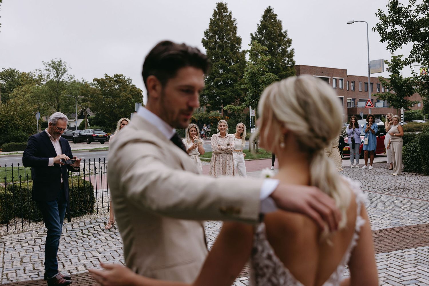 Dancing couple in formal attire on cobblestone plaza with wedding guests in background.
