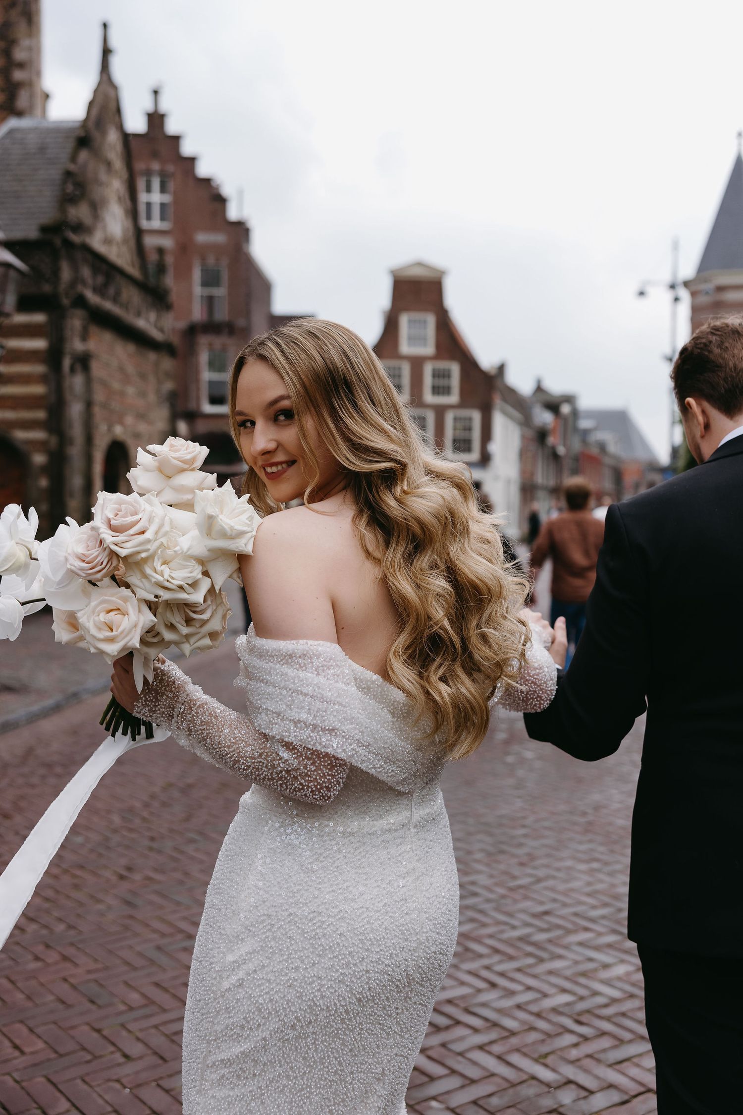 Bride in fitted white dress holds bouquet while walking through historic streets.
