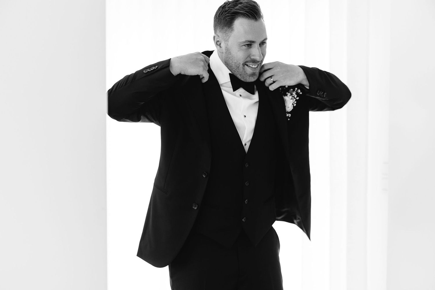 A groom in black tuxedo and bowtie adjusts his jacket.